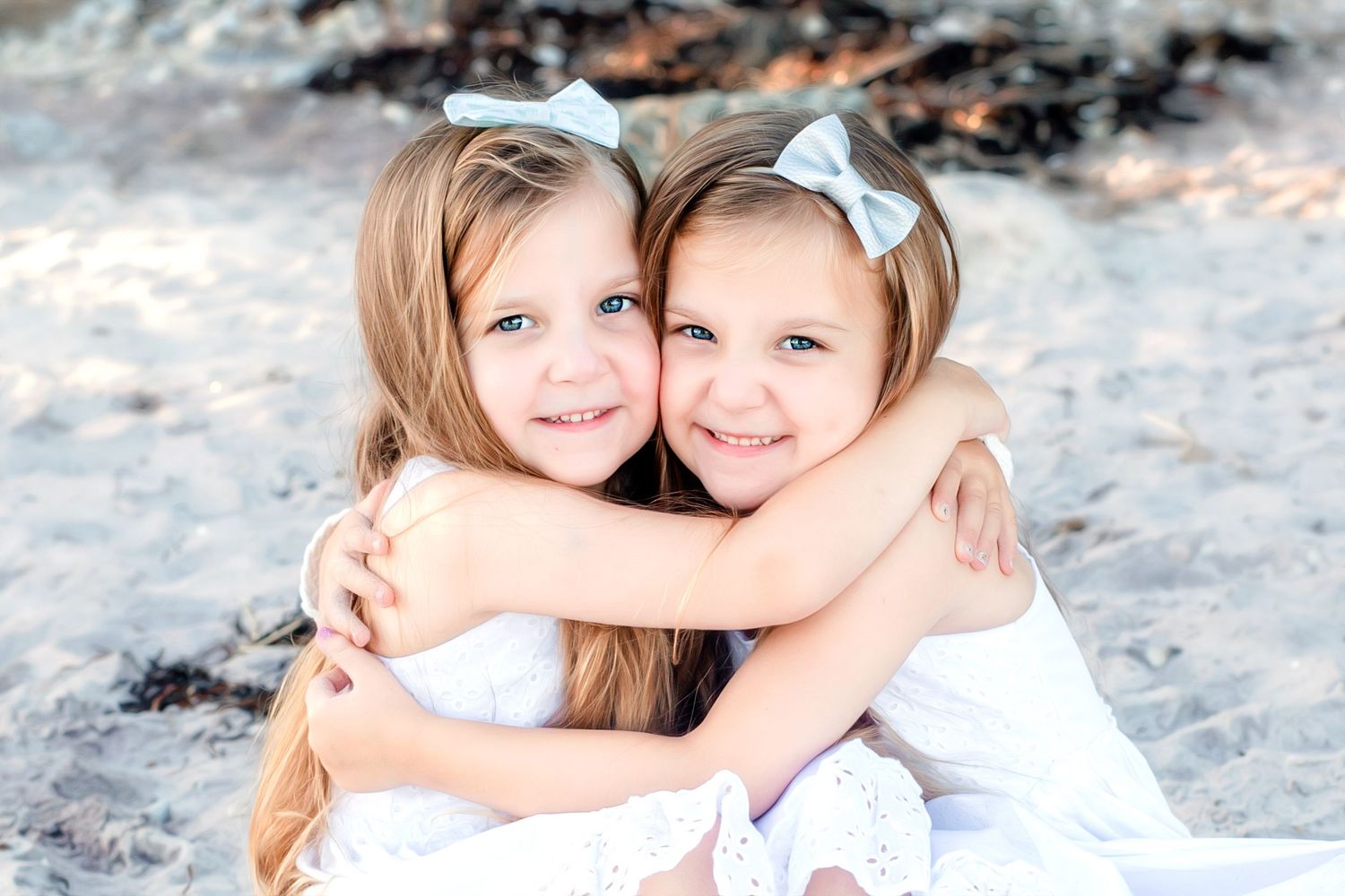 Two young children in white outfits embrace and smile while sitting together outdoors in a natural setting.
