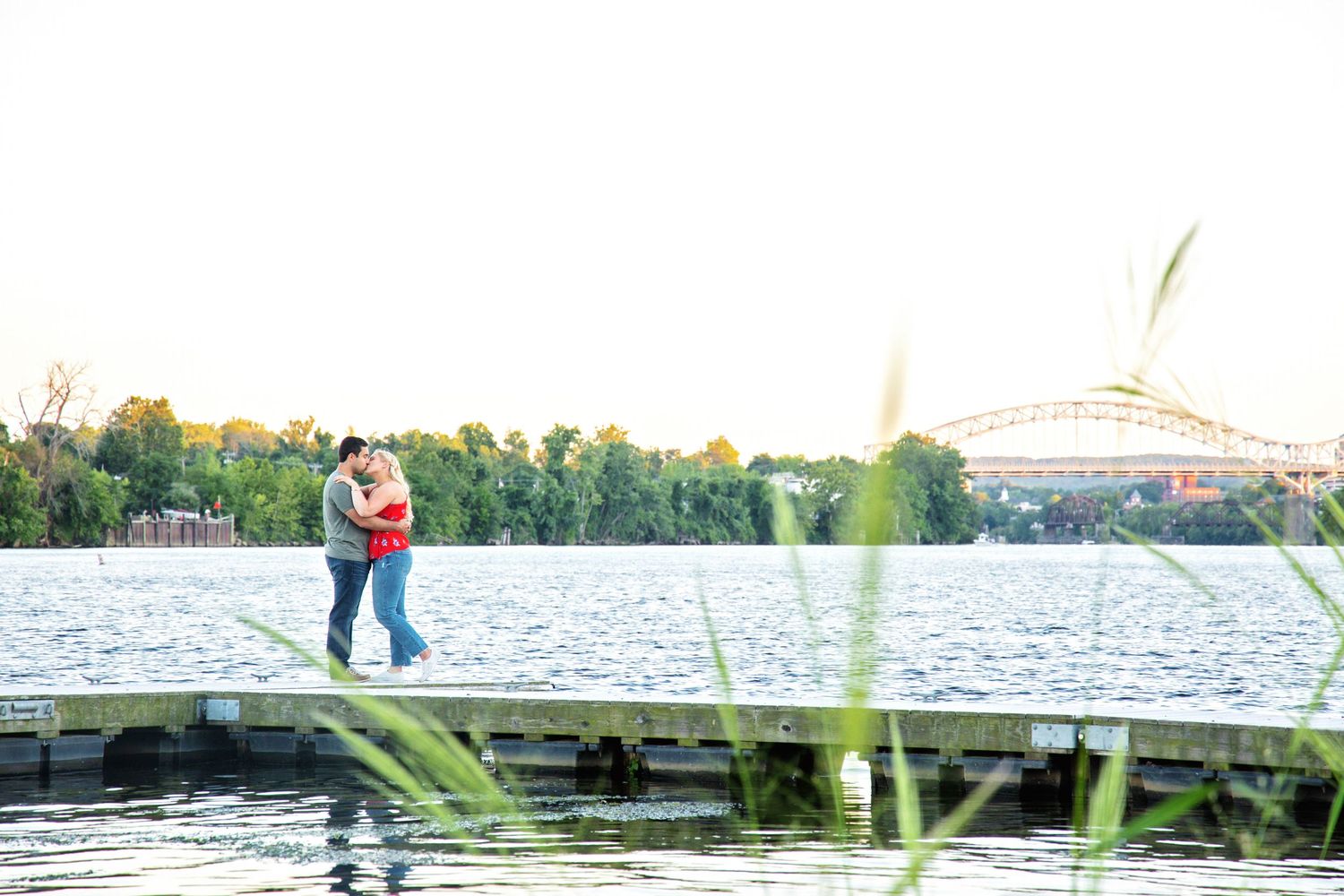 A couple embraces on a wooden dock overlooking a lake with trees and a building in the background.
