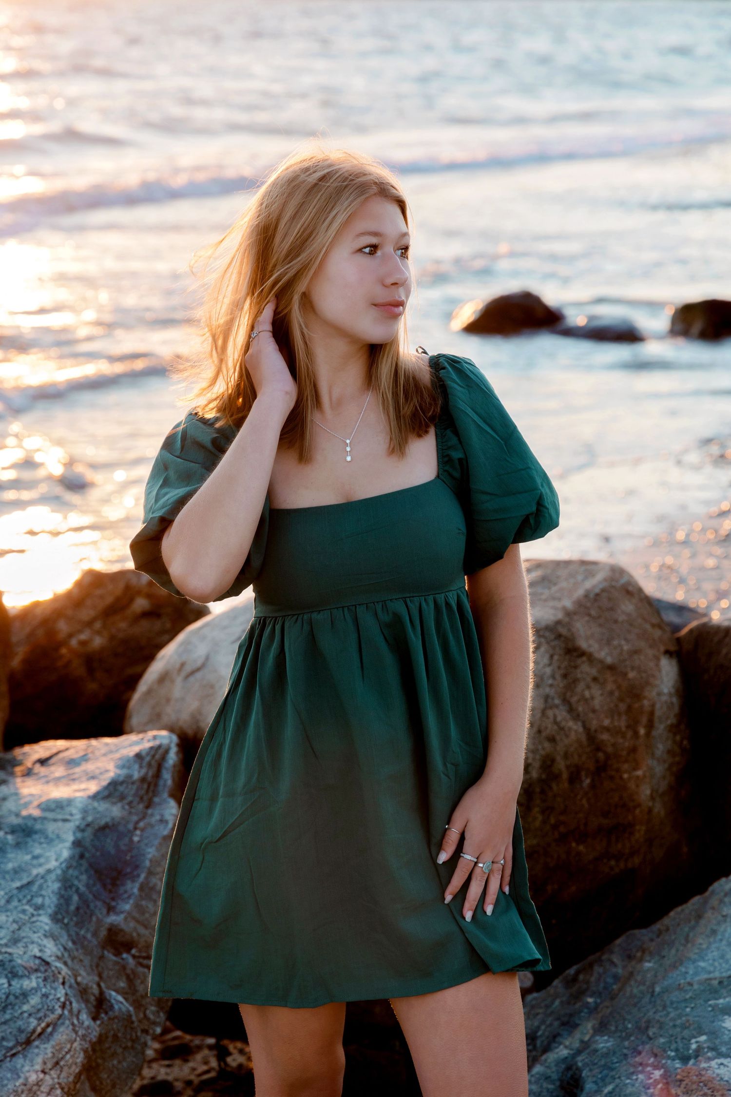 Woman in dark green dress poses gracefully on coastal rocks during golden sunset.