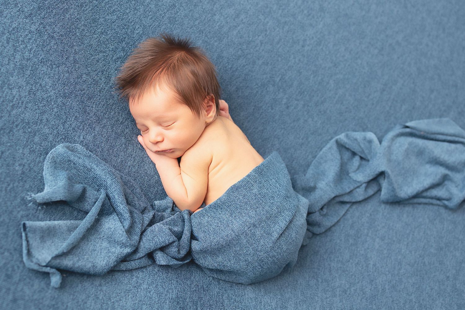 Artistic newborn portrait with baby wrapped in blue fabric against matching background.