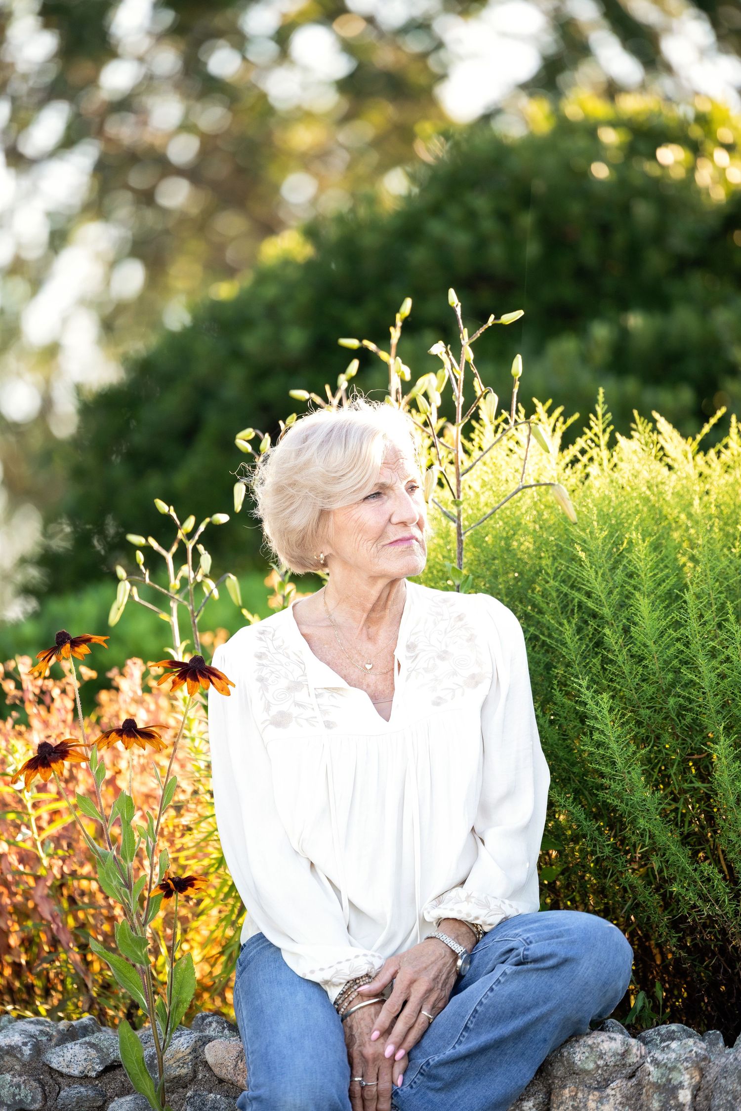 A person in a white shirt and jeans sits on a stone wall at sunset in a garden with blooming flowers and greenery.