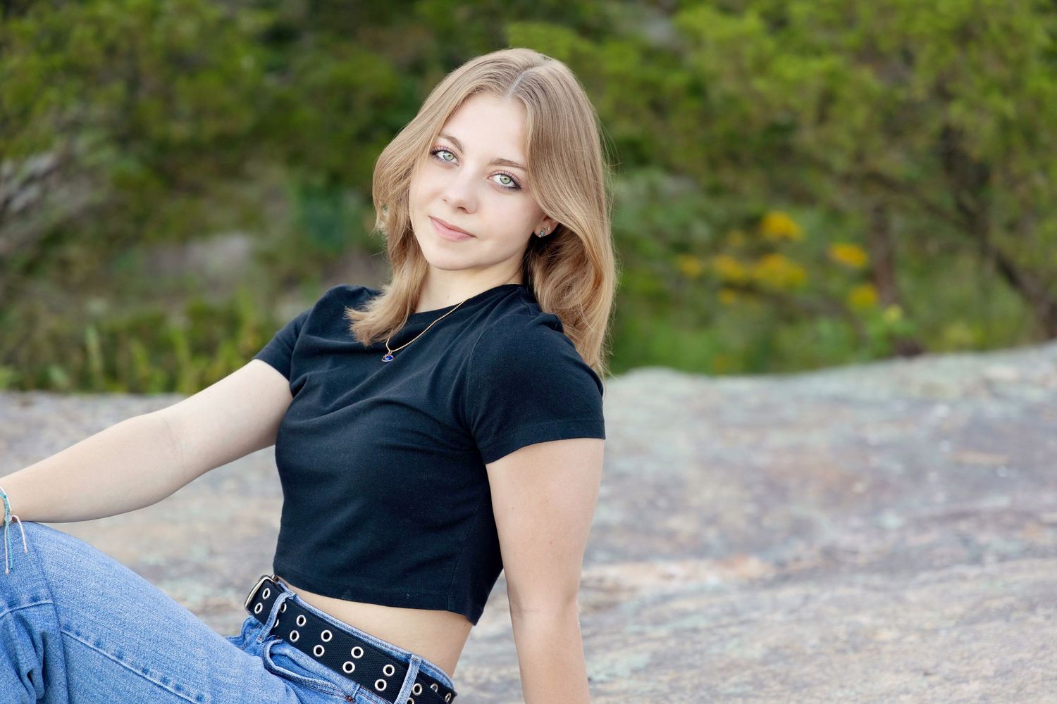 A person in casual attire sits on rocks near a scenic waterfall surrounded by lush greenery and natural scenery.