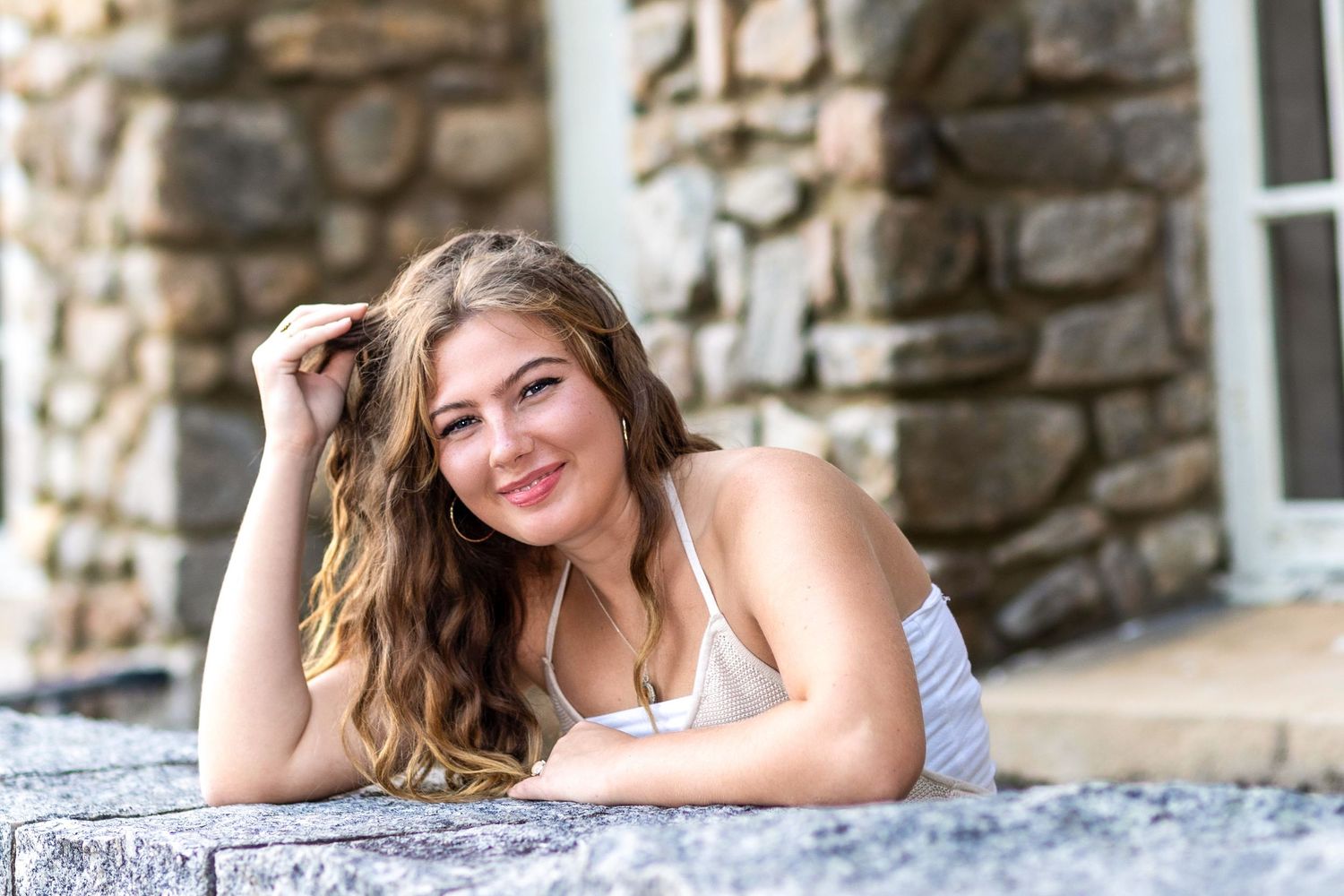 A person in a white dress poses outdoors against a rustic stone wall during a summer photo session.