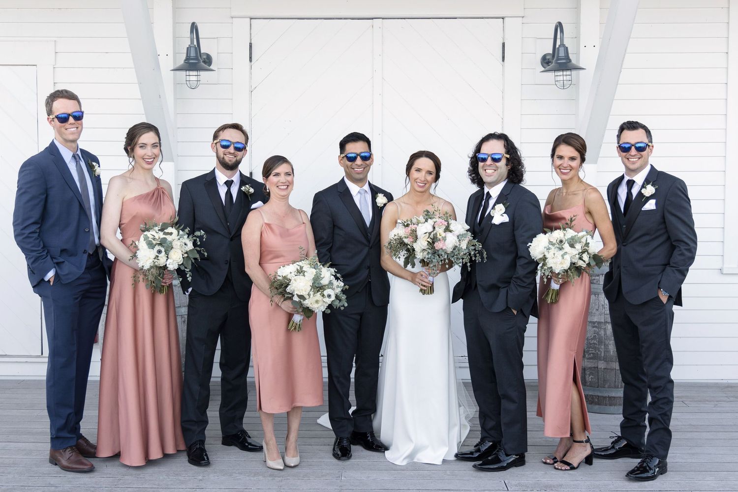 A wedding party poses together in formal attire with bridesmaids in pink dresses and groomsmen in dark suits.