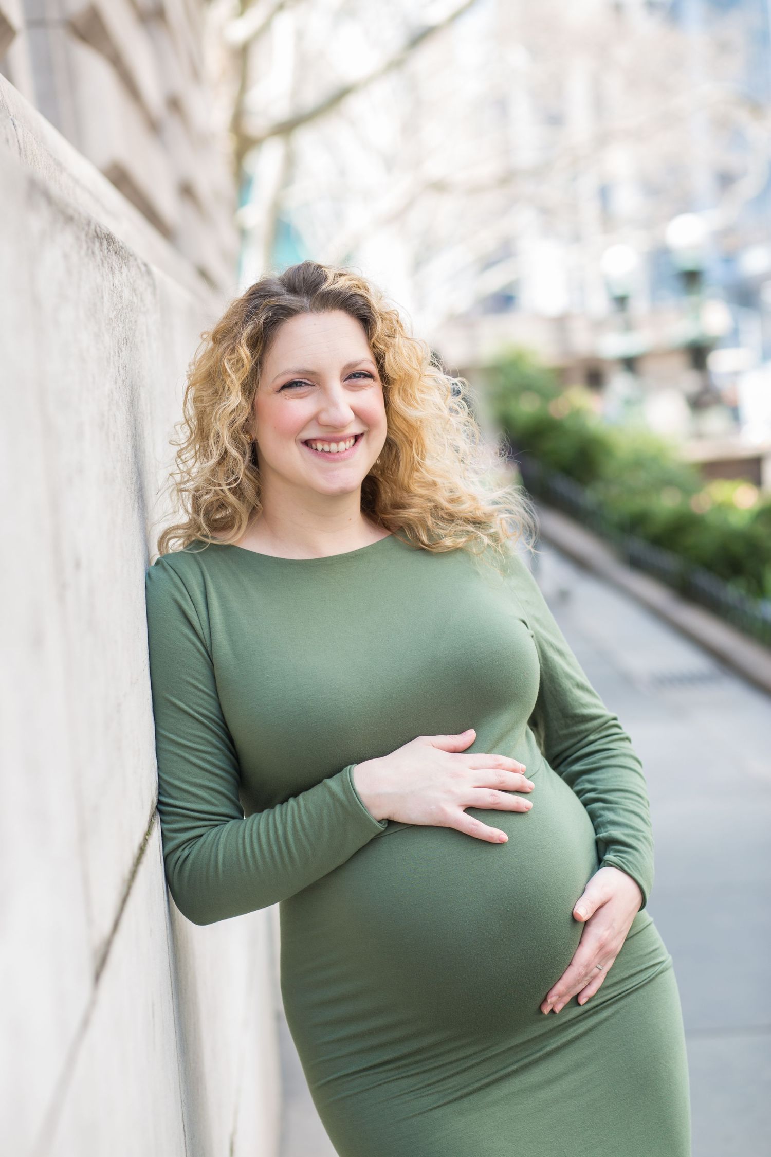 A pregnant woman in a green fitted dress stands against a light wall outdoors on a sunny day.