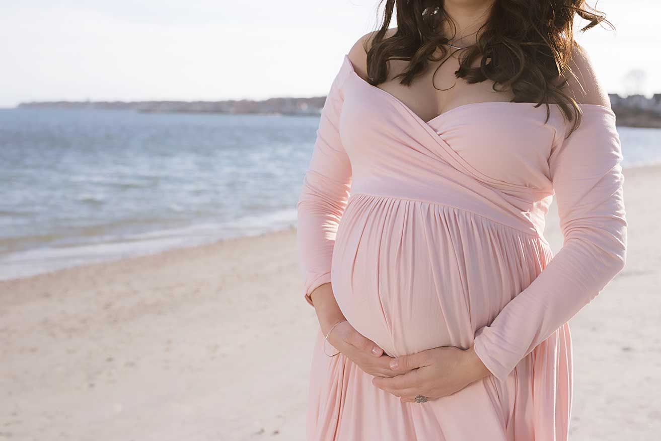 A pregnant woman in a soft pink dress stands on a beach at sunset, cradling her baby bump.