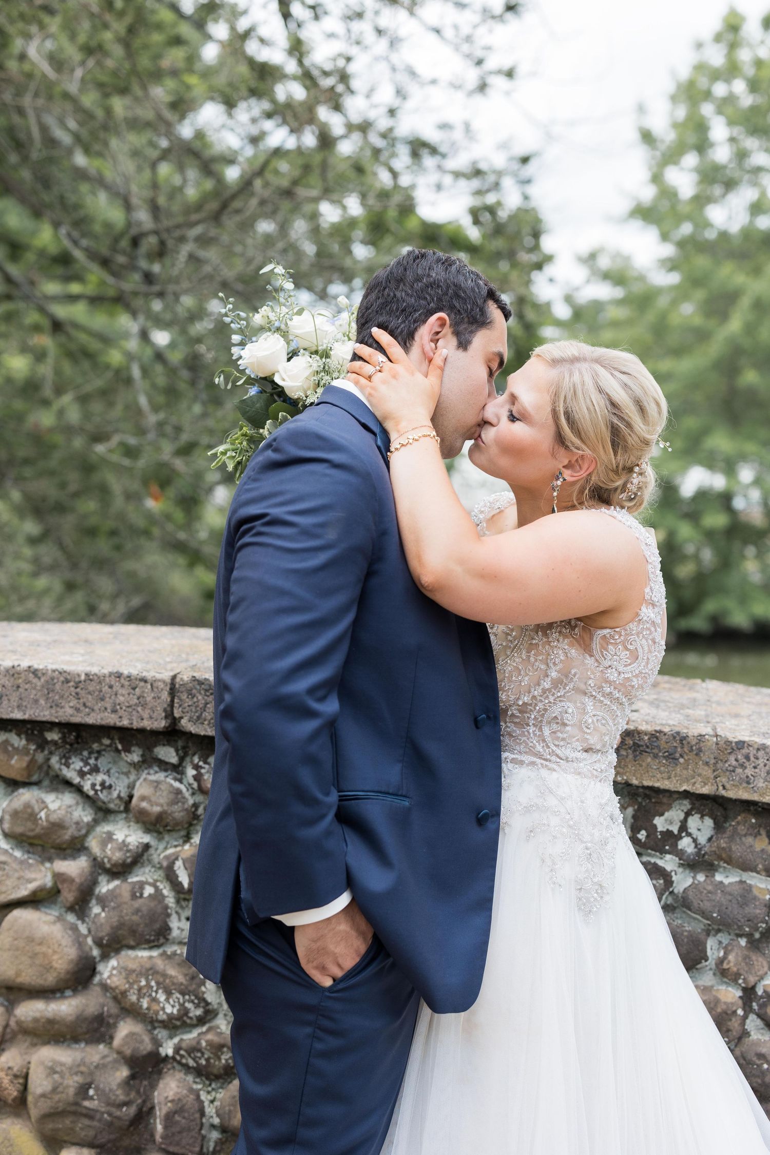 A romantic wedding couple shares intimate kisses in front of a rustic stone wall in an outdoor garden setting.