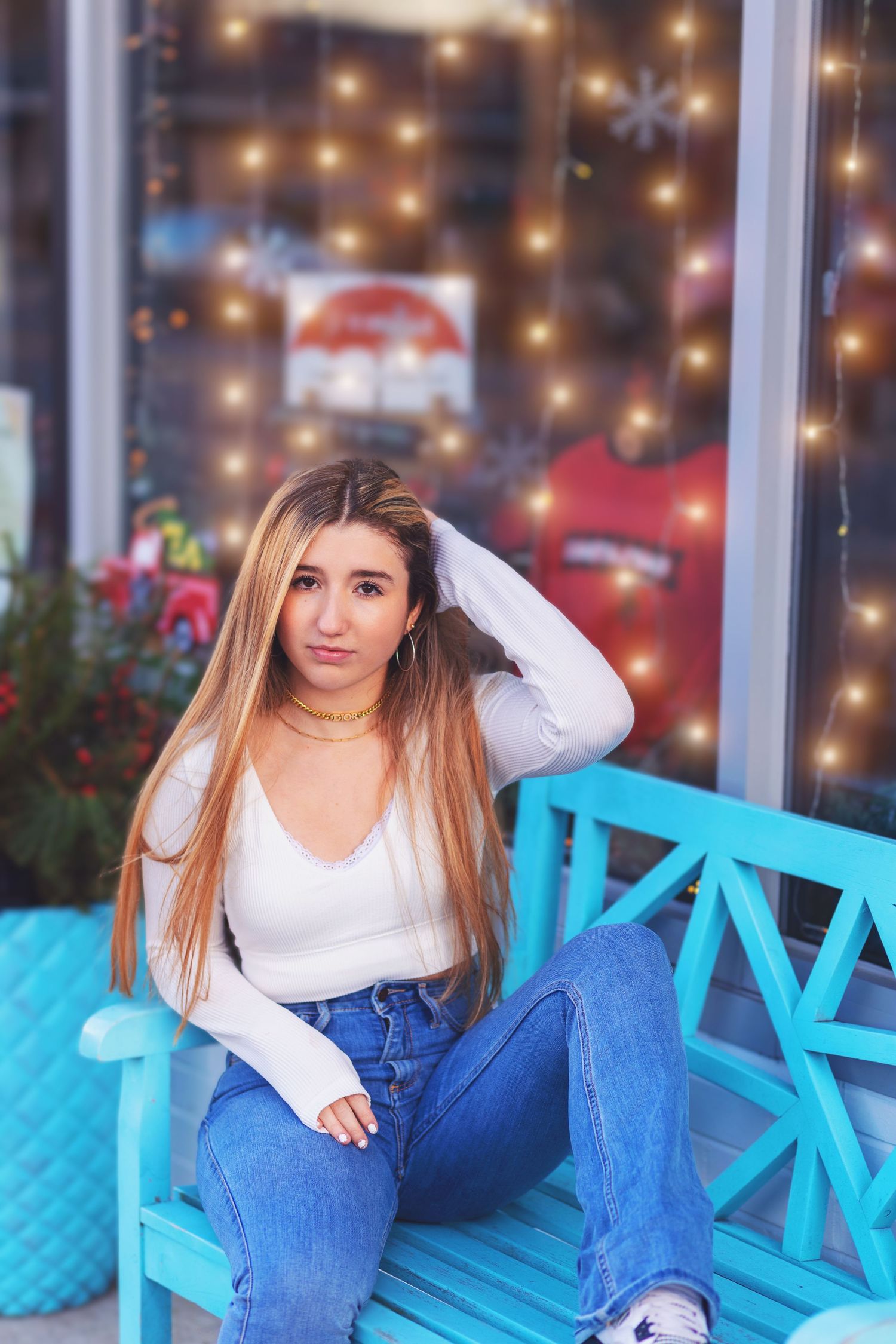 Person in white top and jeans sits on bright turquoise bench with twinkle lights and holiday decor in background.