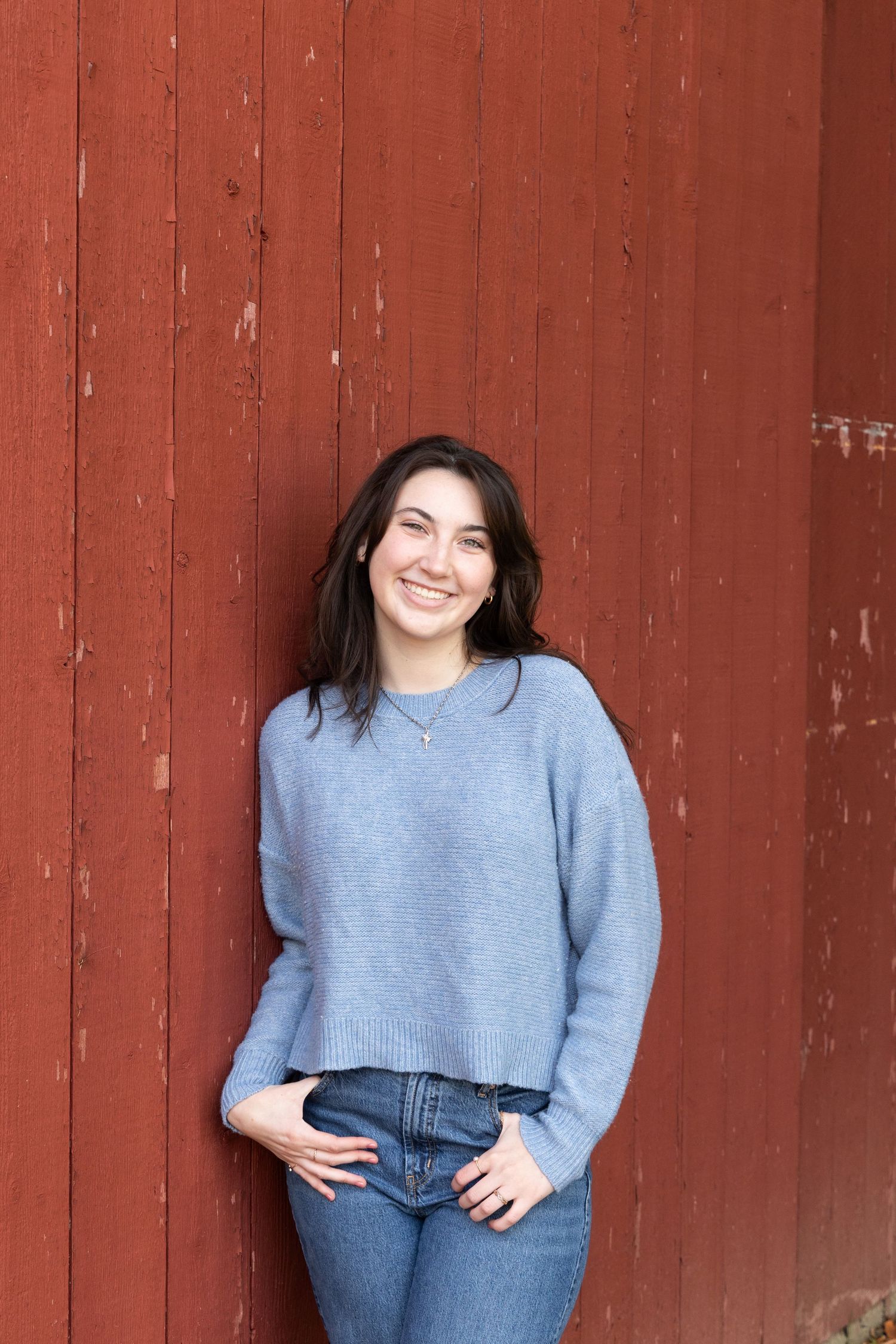 Casual portrait session against a rustic red barn wall wearing blue sweater and jeans.
