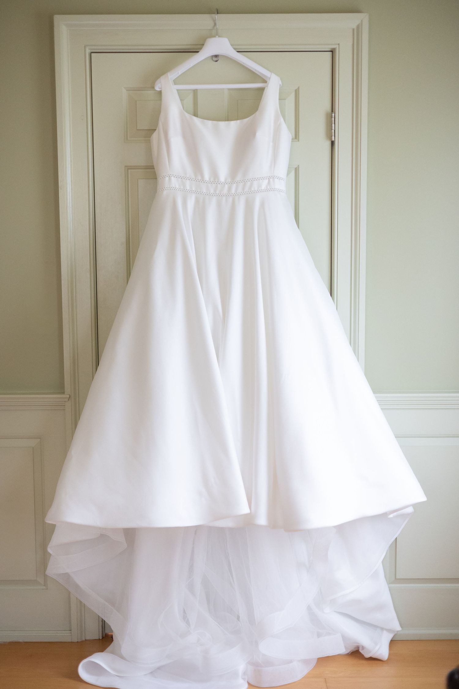 Elegant white wedding dress with full skirt hanging on display against cream colored door.