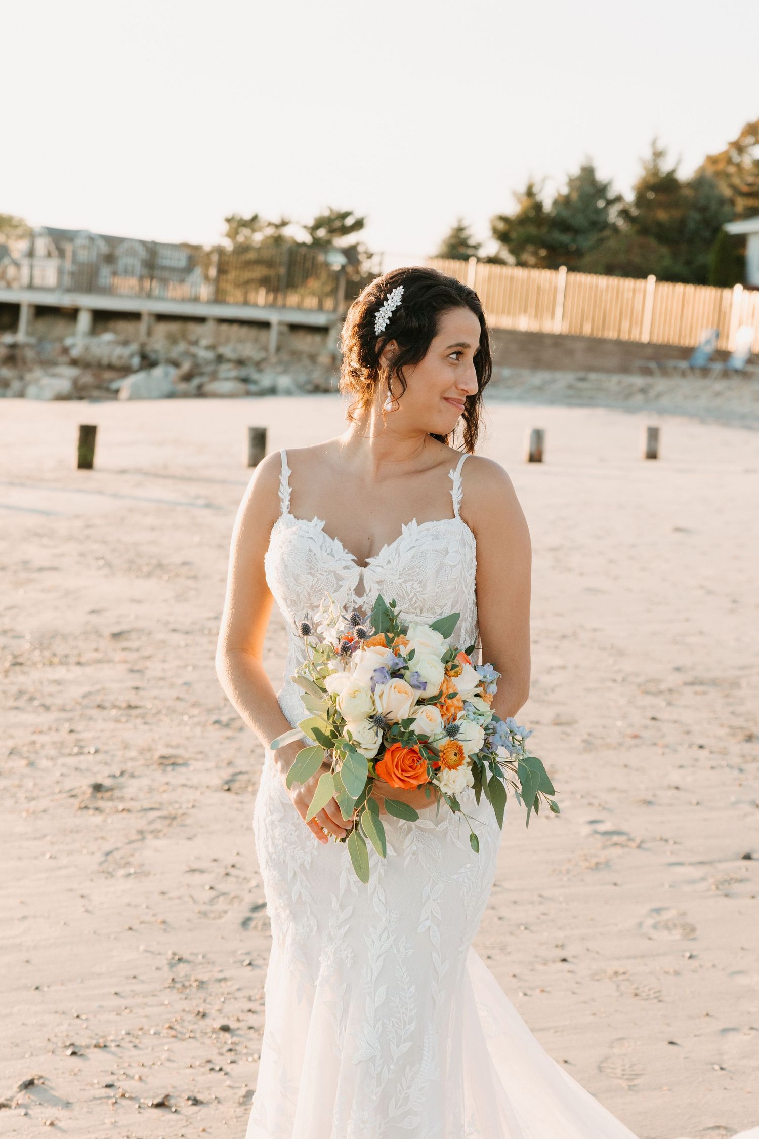 Bride in lace gown holding vibrant orange and white bouquet on sandy beach.