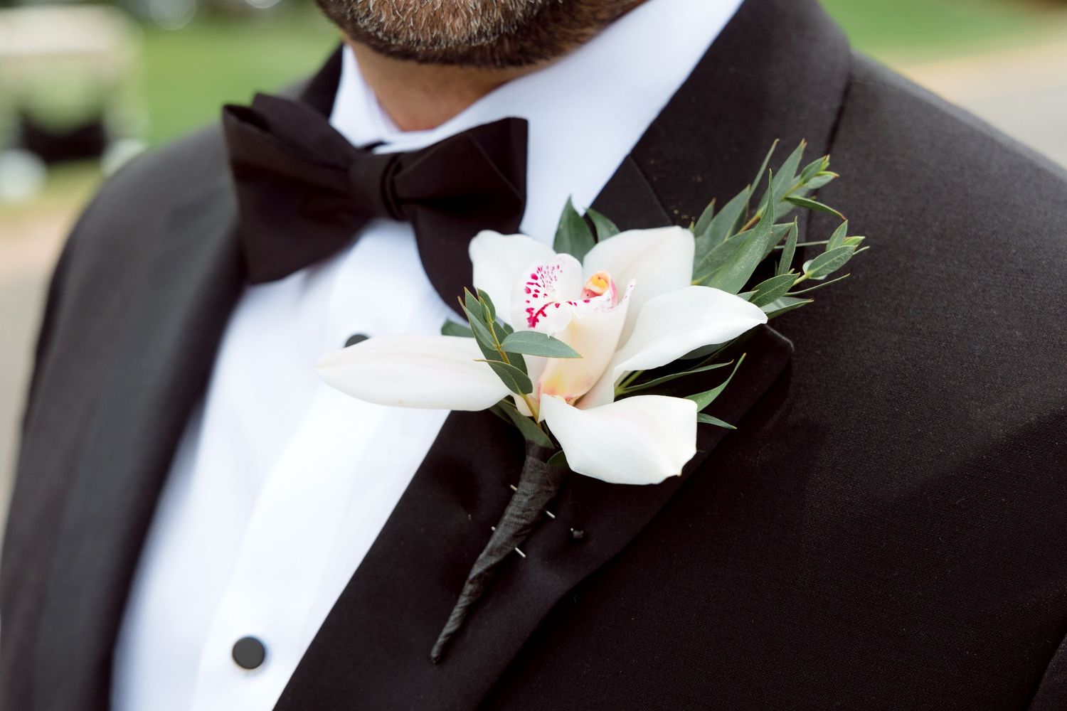A black tuxedo lapel with a white rose and greenery boutonniere pinned to it.