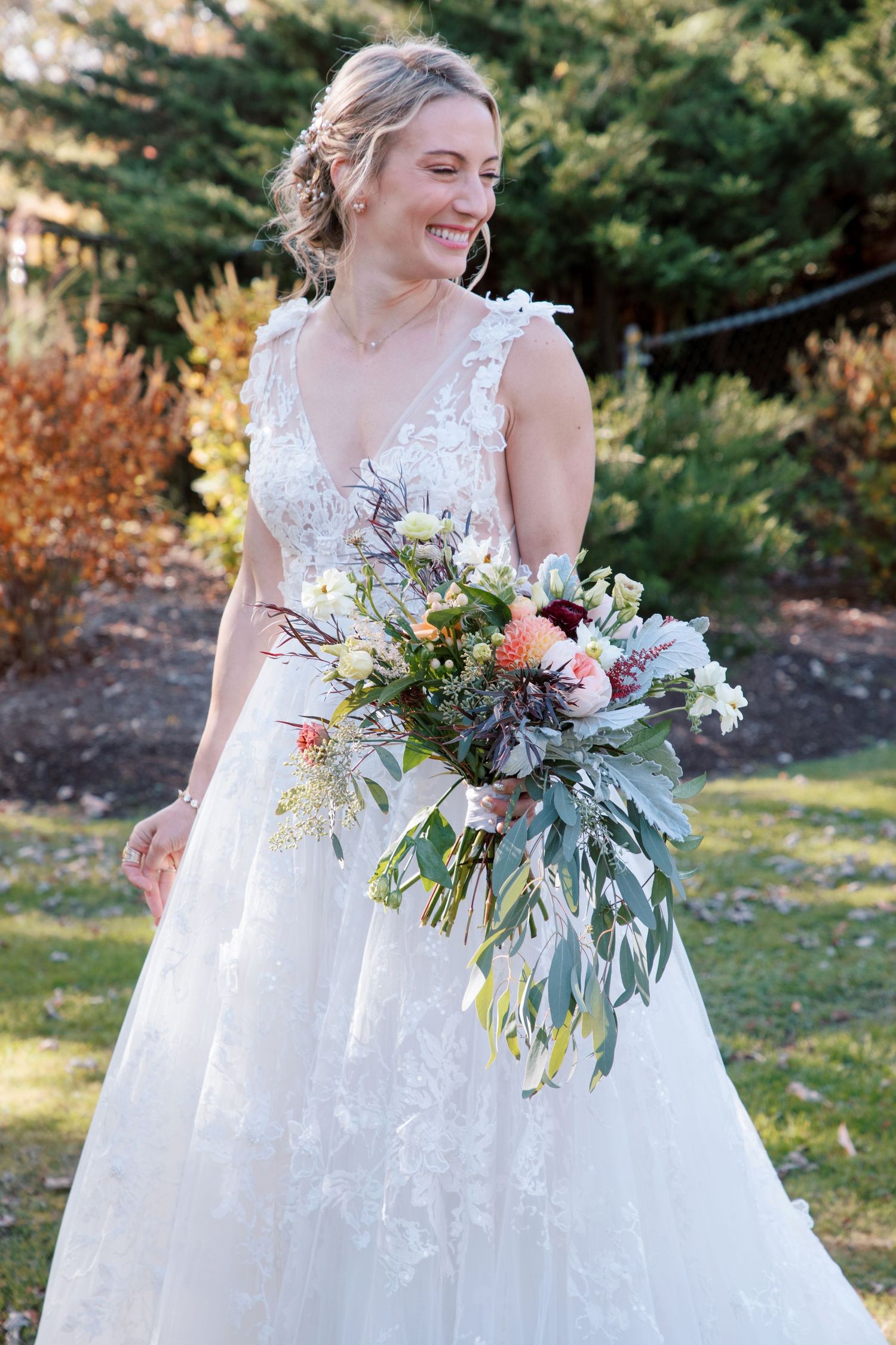 A bride in a white lace wedding gown holds a beautiful bouquet of mixed flowers and greenery against an outdoor autumn backdrop.