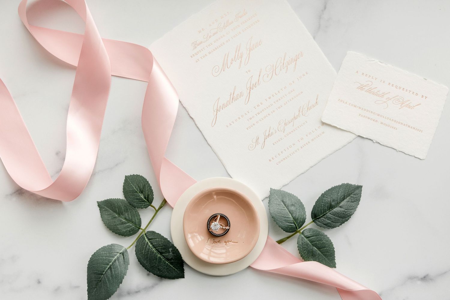 Pink silk ribbon and green eucalyptus leaves arranged with wedding stationery on white marble background.