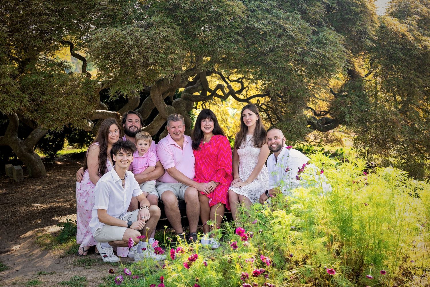 A group poses together outdoors in coordinated pink and white outfits among flowering garden foliage.