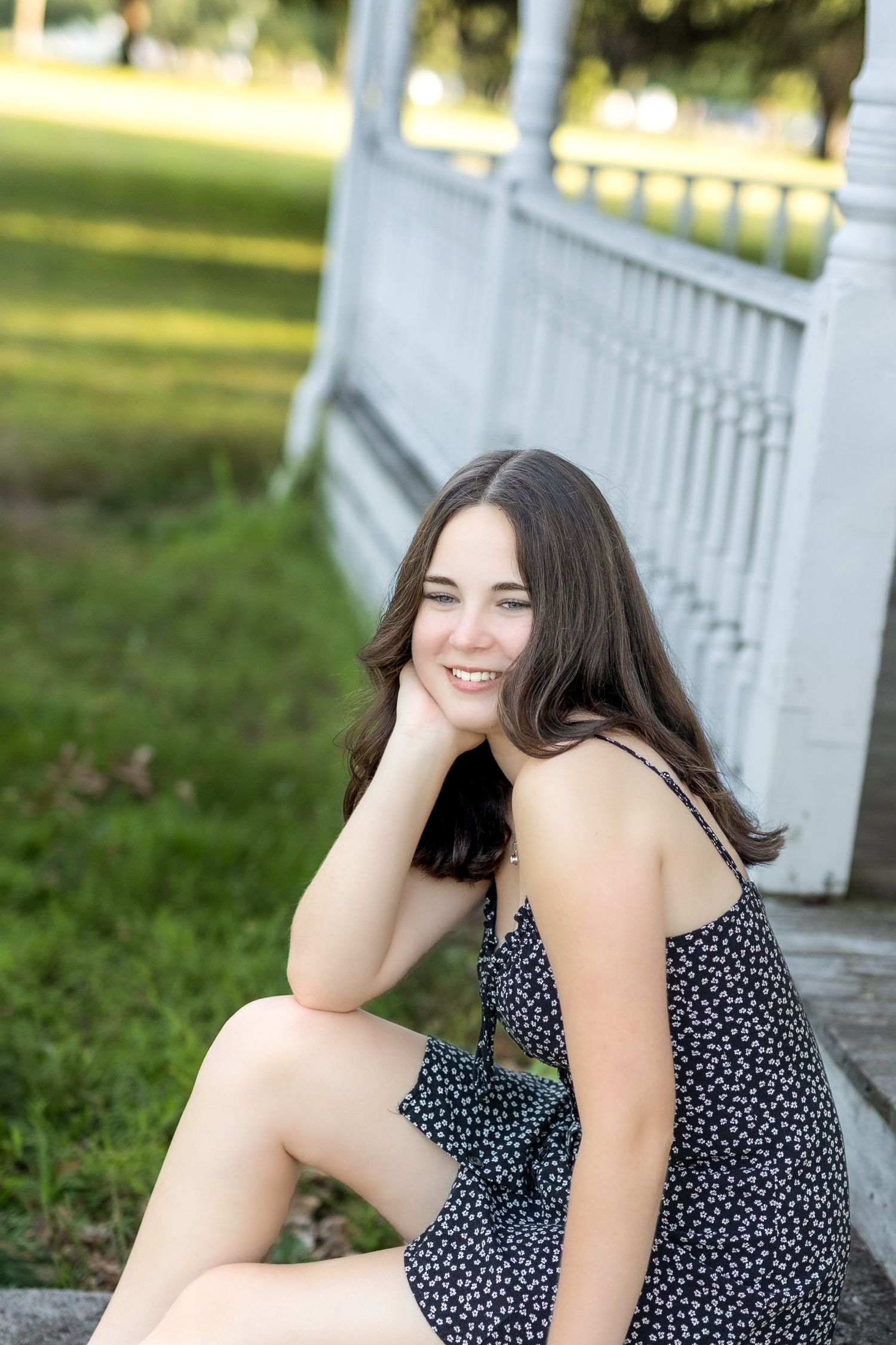 A person in a polka dot dress sits and poses outdoors near a white railing on a sunny day.