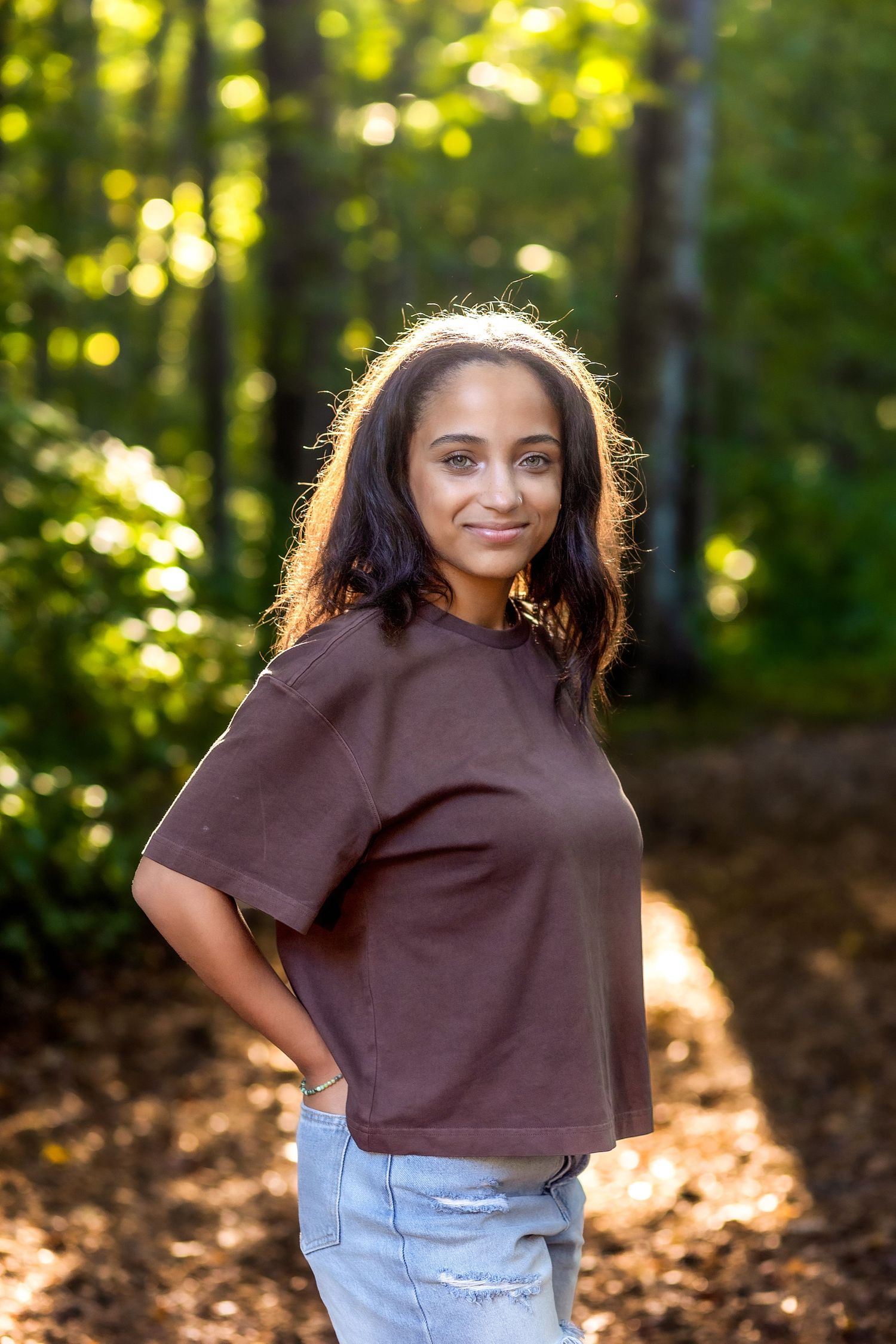A person in a brown crop top and light jeans poses outdoors in a sunlit forest setting.