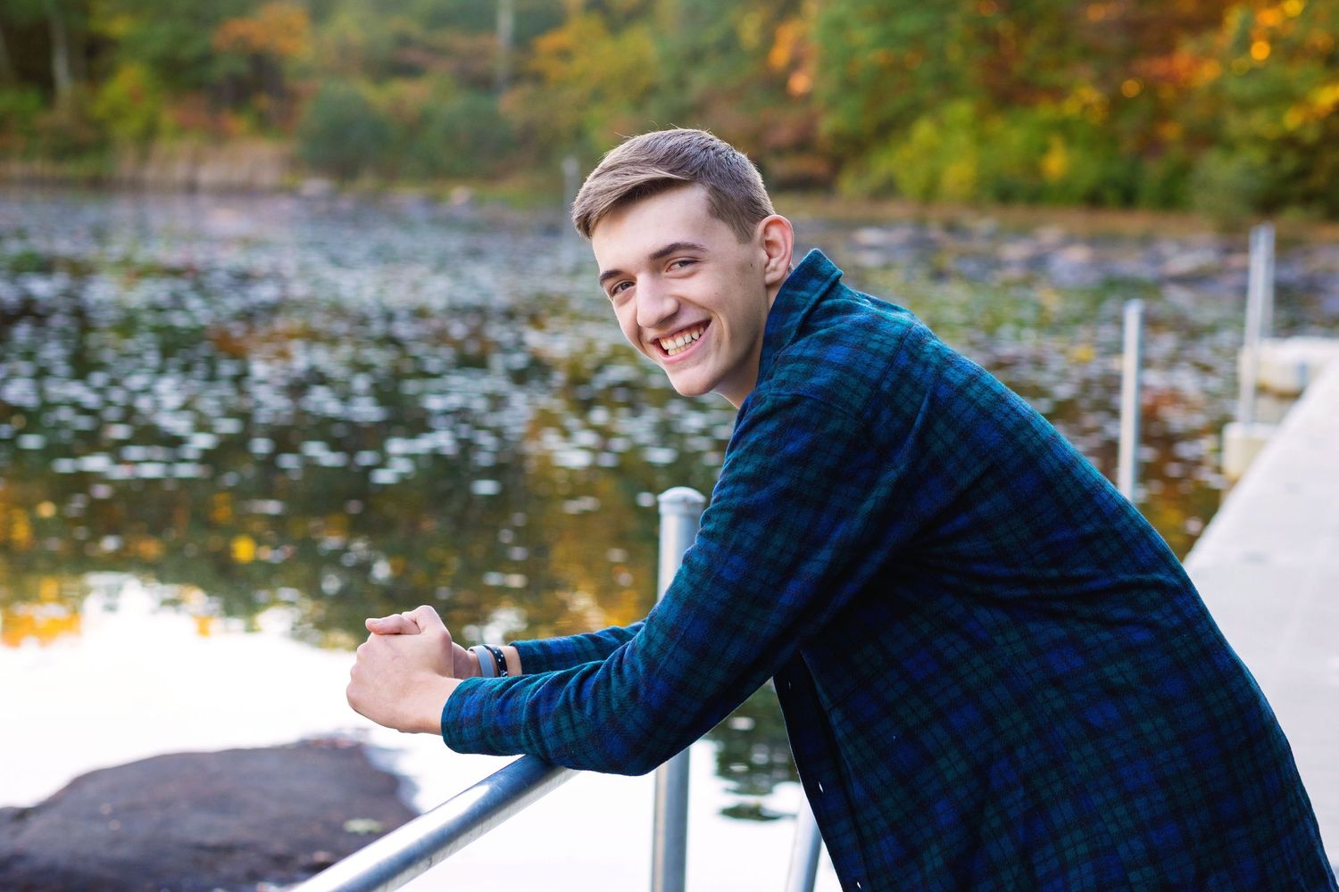 A person in a blue sweater poses near a scenic pond with autumn foliage in the background.