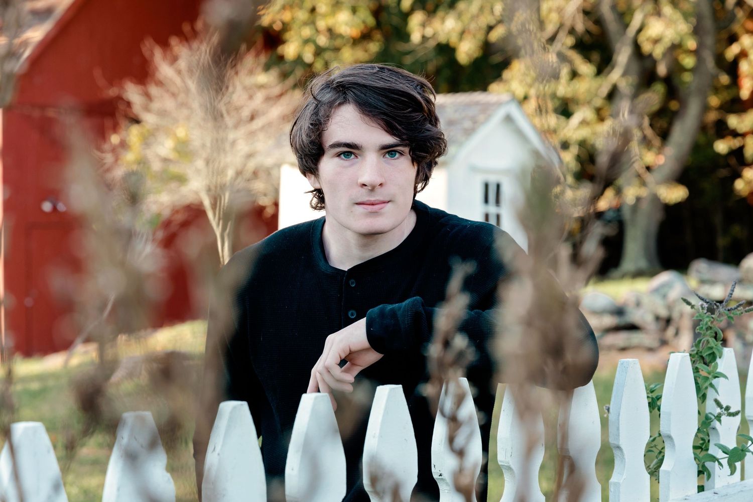 A person stands behind a white picket fence on a sunny autumn day with trees in the background.