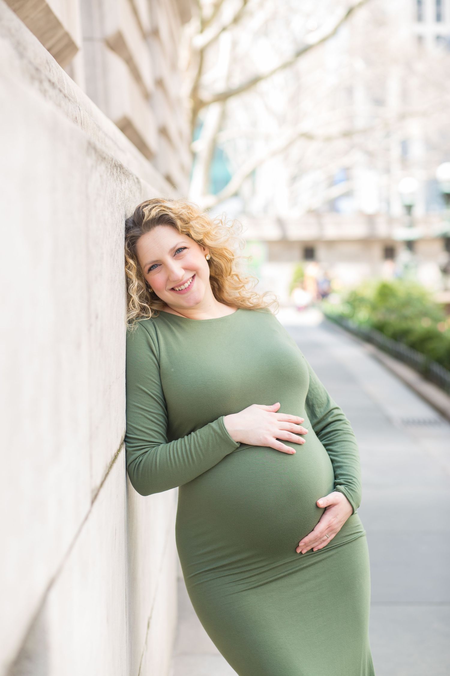 A person in a fitted green long sleeve maternity dress poses against a light colored wall outdoors.