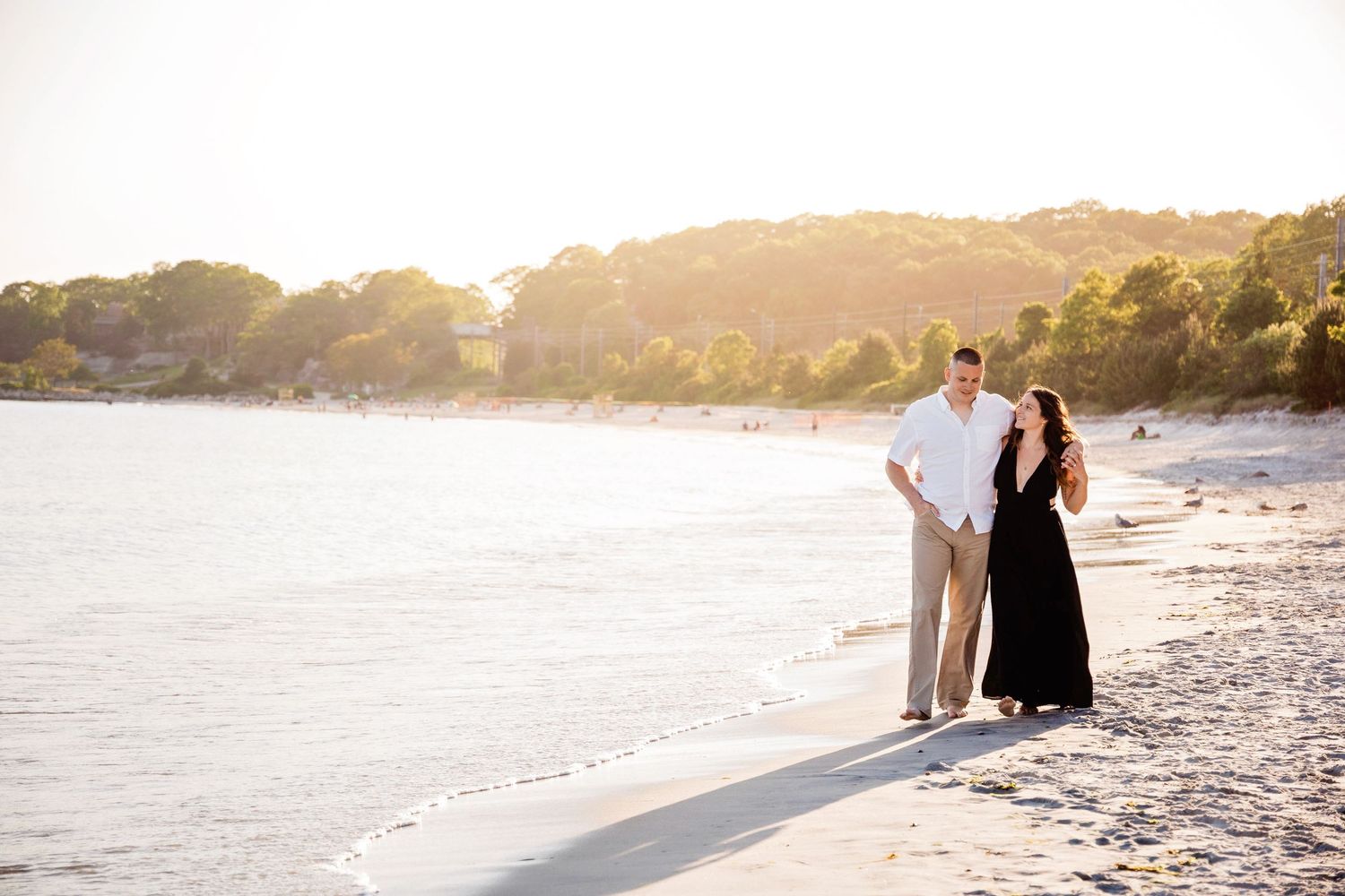 A couple walks along a sandy beach at sunset with golden light illuminating the coastline.