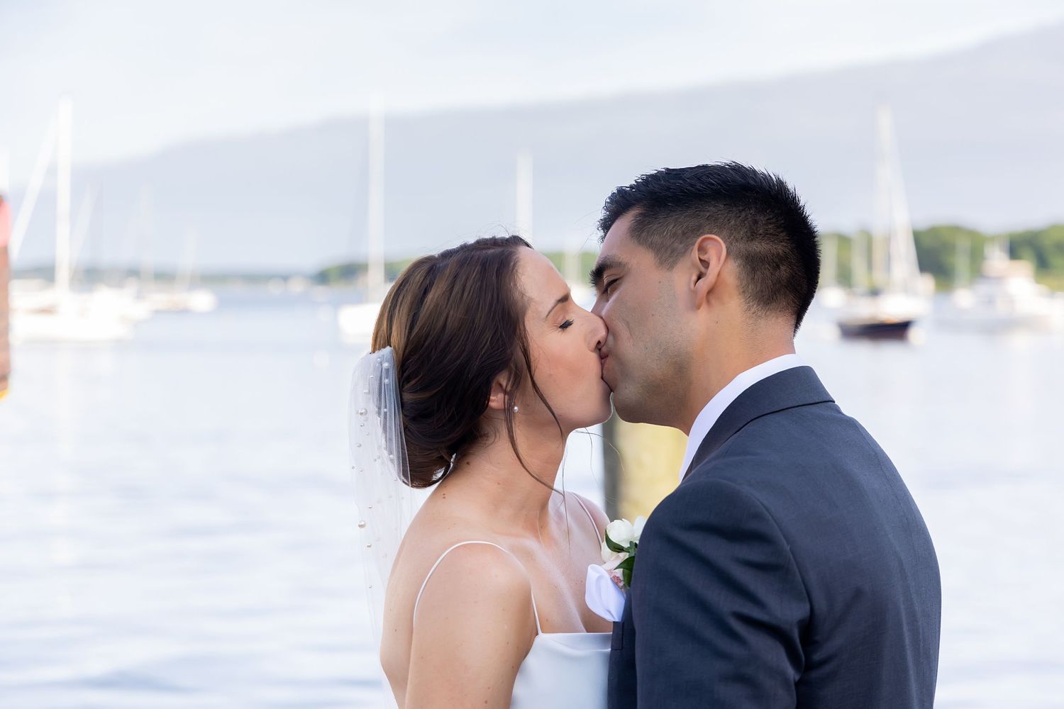 A romantic couple shares a tender kiss on a dock with boats and water in the background.
