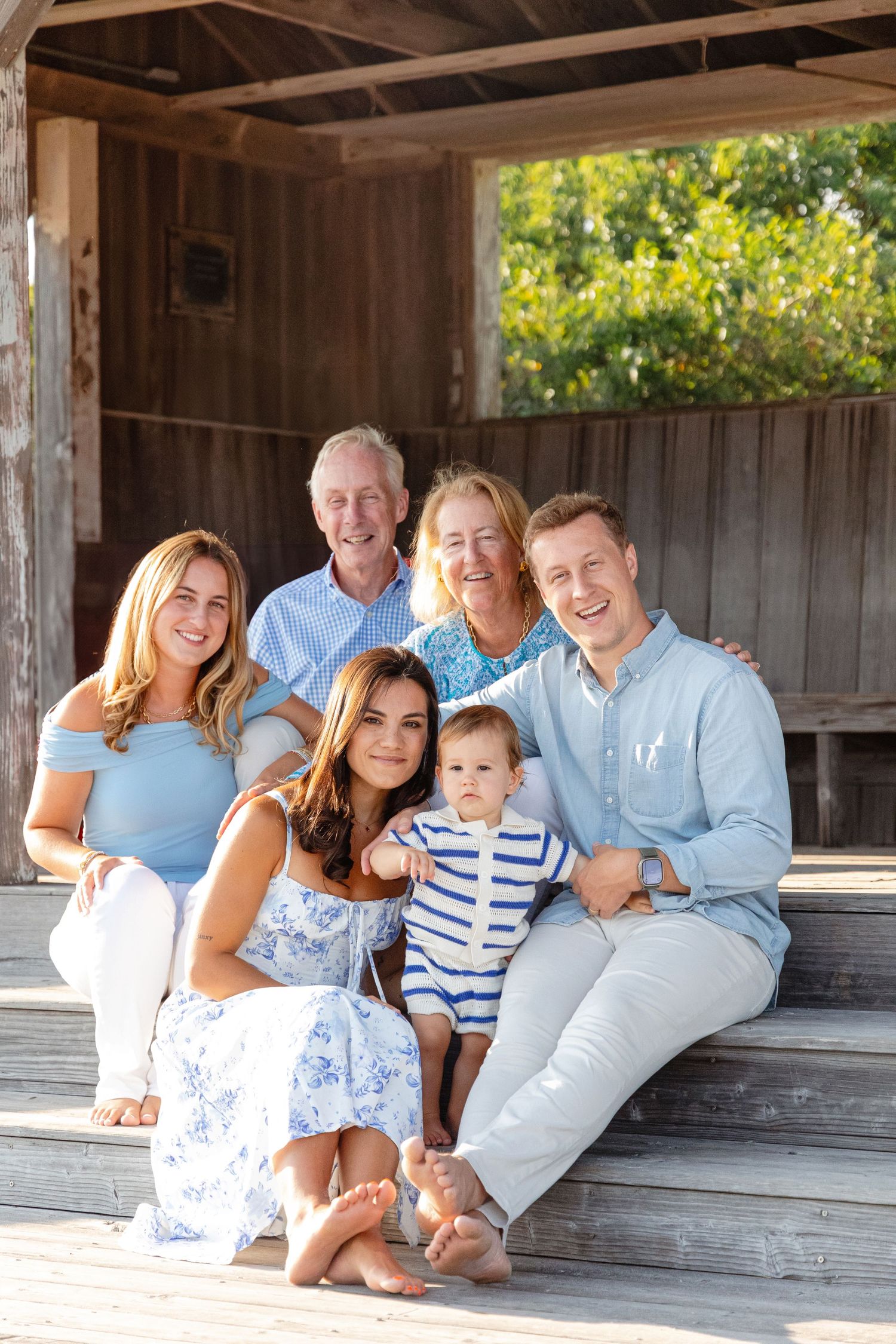 A family wearing coordinated blue and white outfits poses together on a rustic wooden porch in summer.