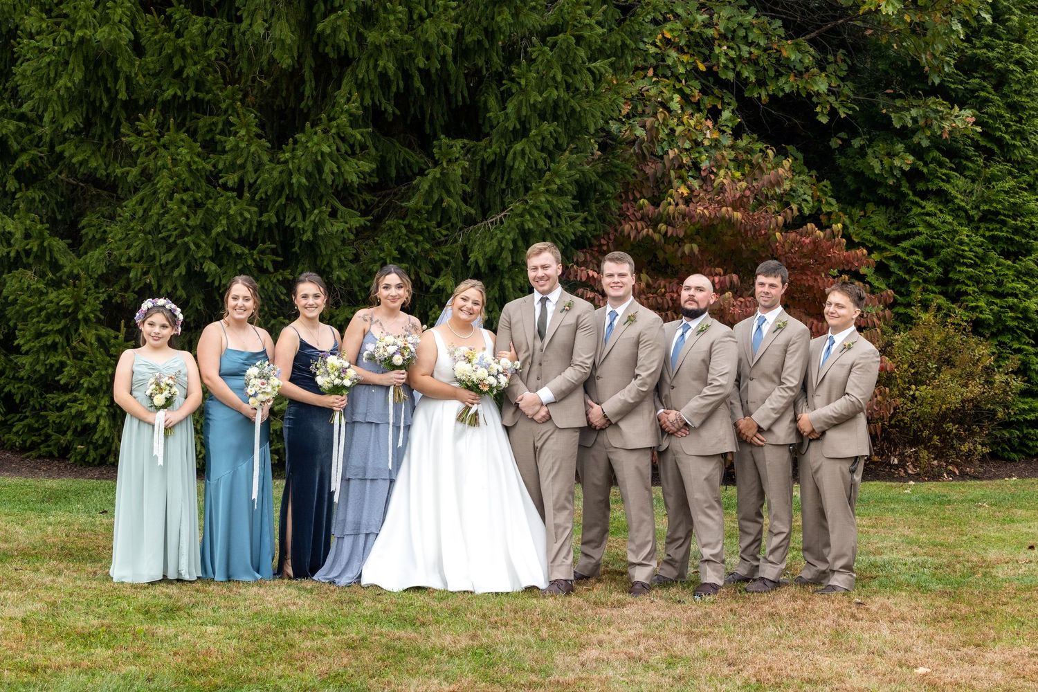 Wedding party in blue dresses and gray suits poses formally on garden lawn.