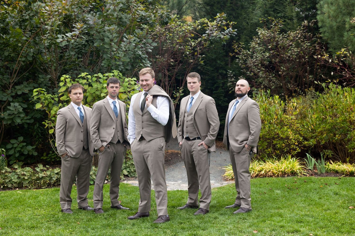 Groomsmen in light gray suits pose together on green lawn with garden backdrop.