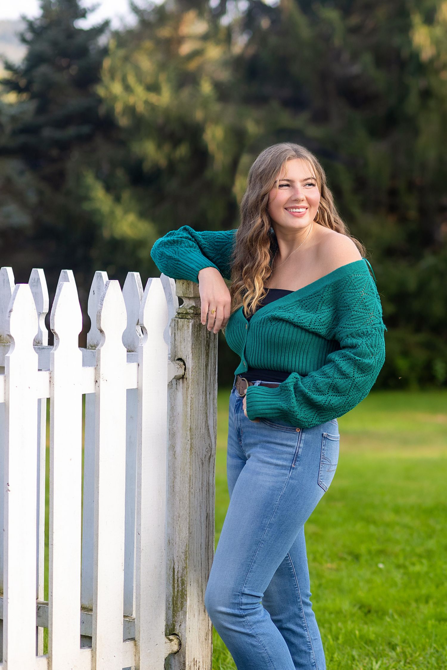 A woman in a green sweater and jeans poses playfully against a white picket fence in a grassy outdoor setting.