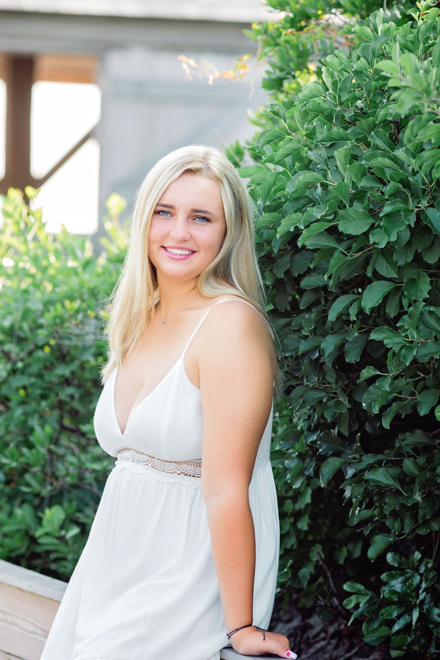 A person in a white sundress stands smiling next to lush green foliage on a sunny day.