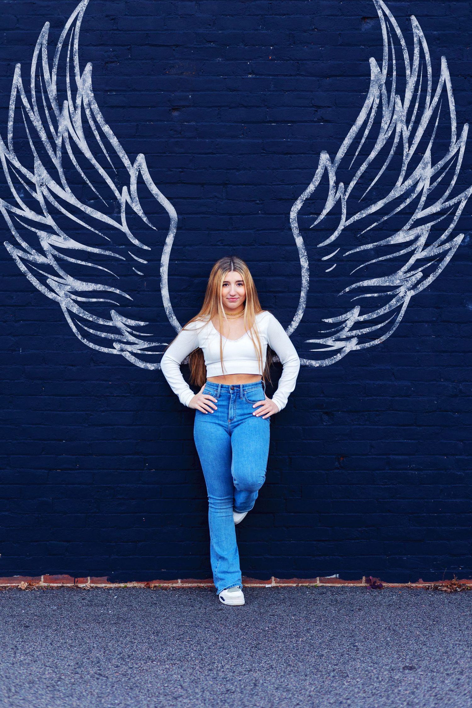 Person poses in front of blue wall with white chalk angel wing art wearing white top and blue jeans.