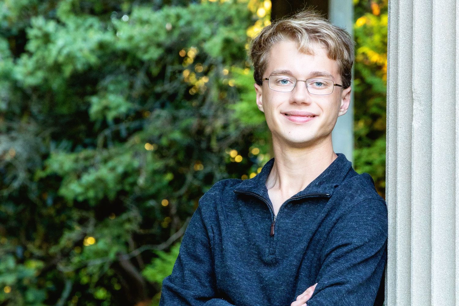 Young person in glasses and dark shirt poses outdoors against green foliage background in natural lighting.
