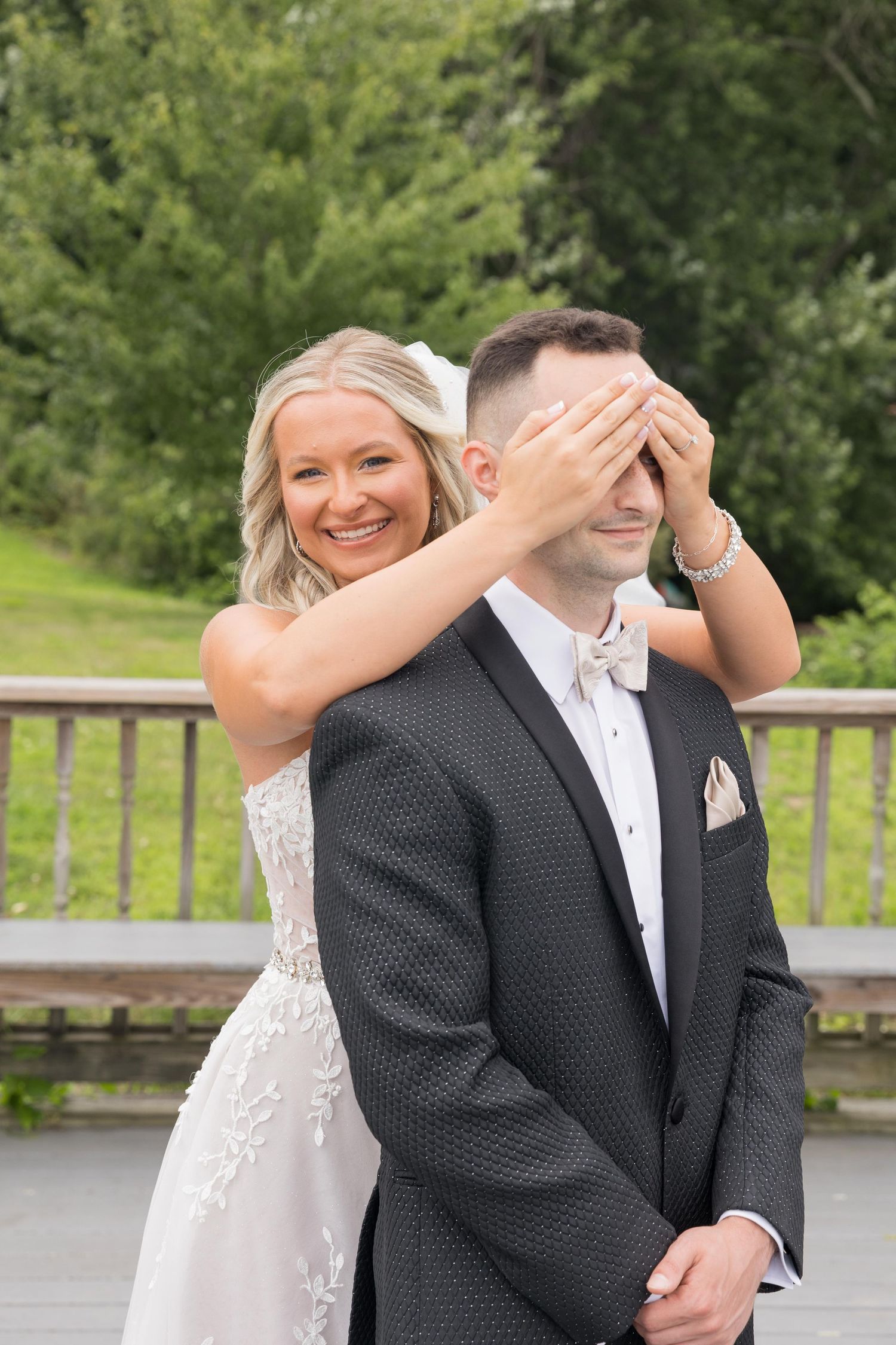 A couple in wedding attire shares a playful first look moment outdoors on a wooden deck.