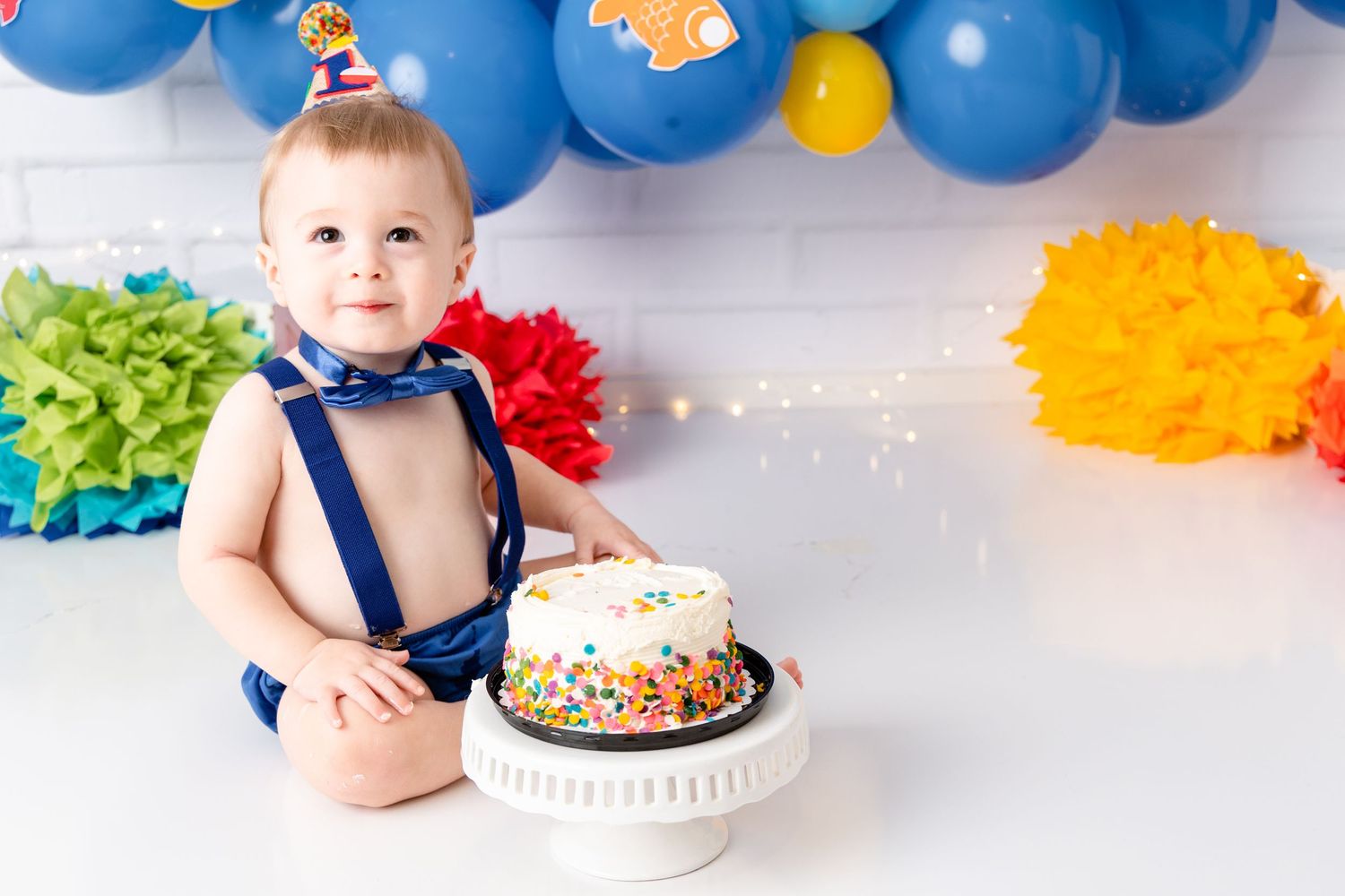 Colorful first birthday cake smash photo session with blue balloon arch and sprinkle decorations.