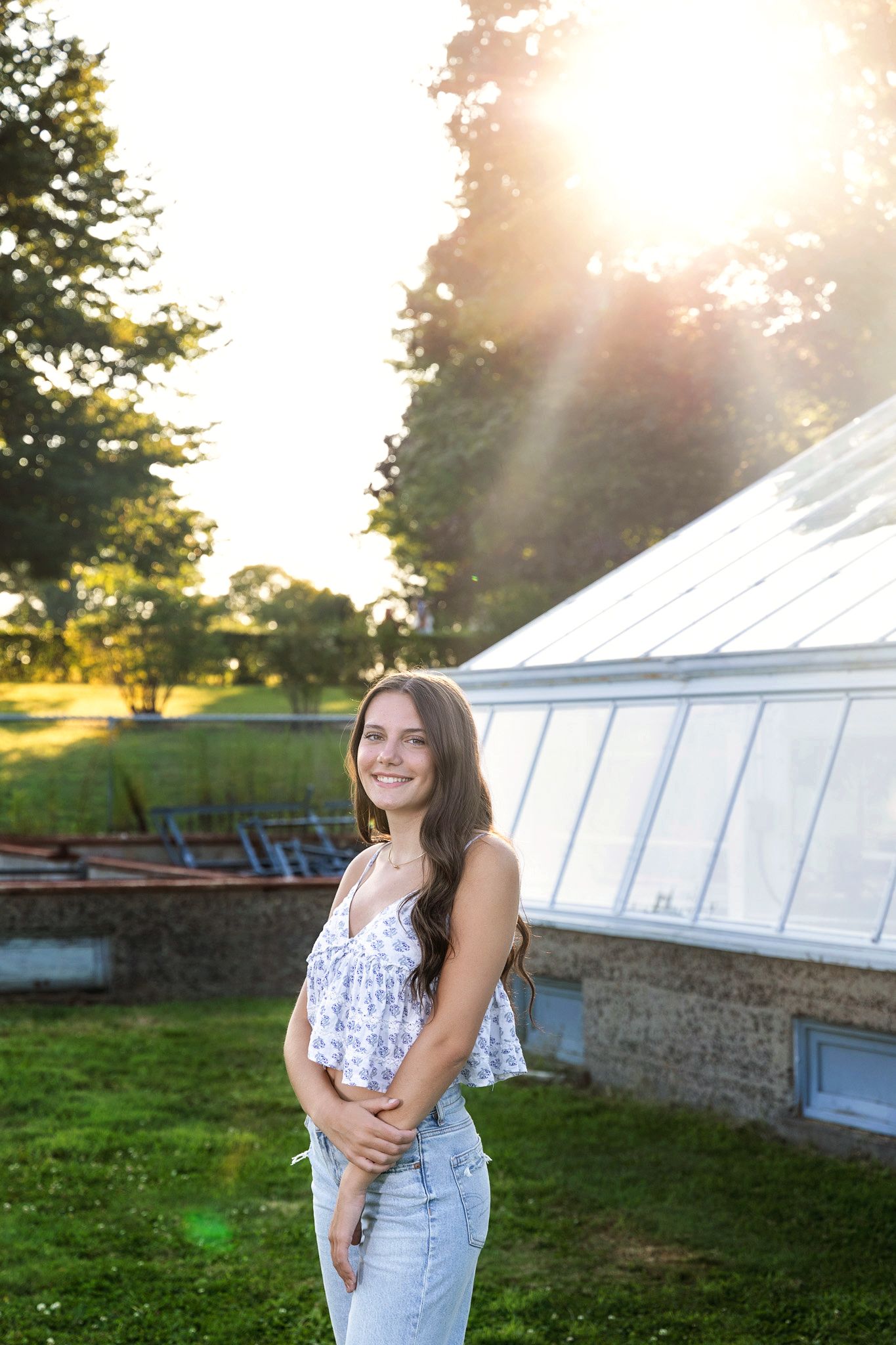 A person in overalls poses and smiles outdoors near a greenhouse structure at sunset.