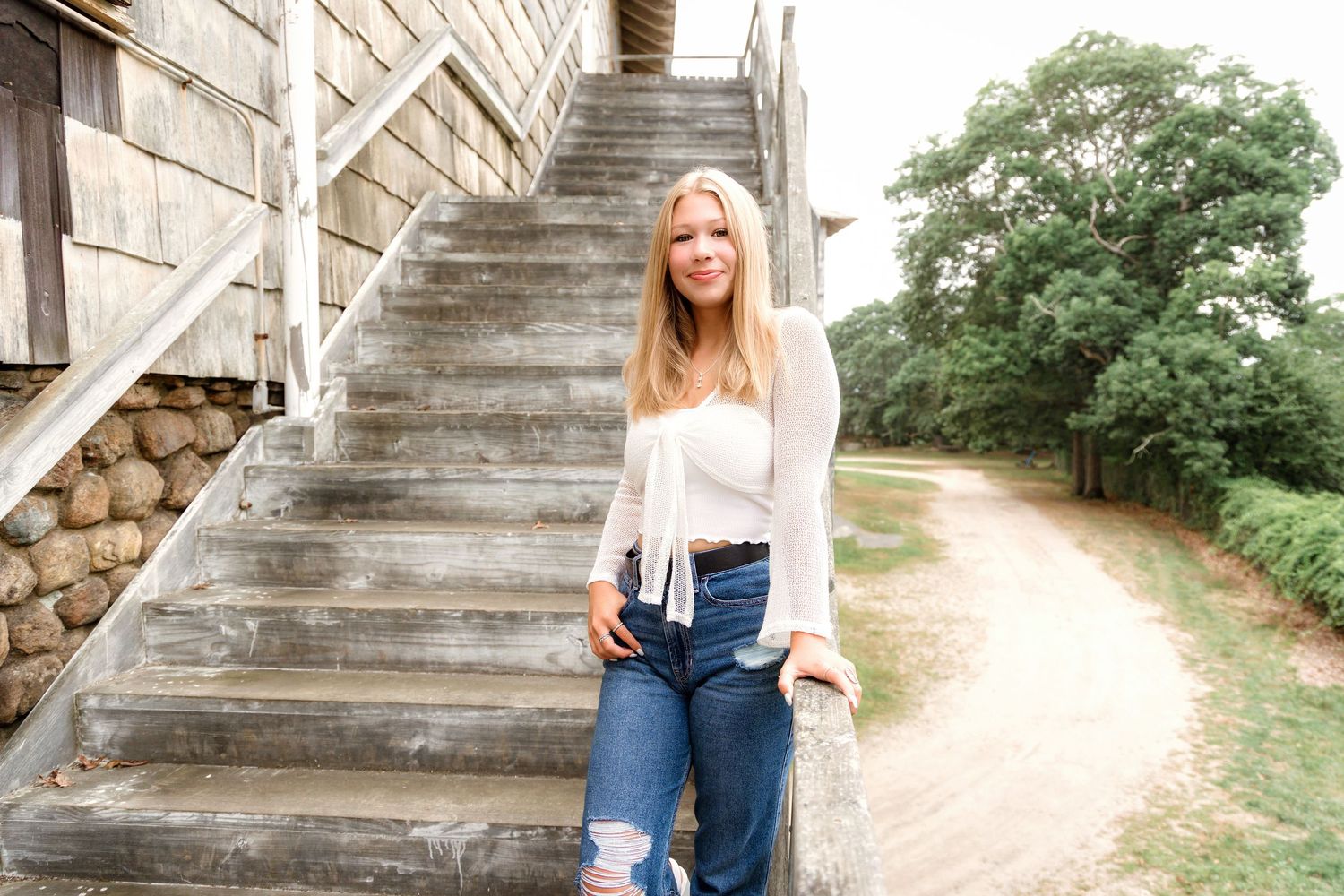 A person in blue jeans and white top sits on rustic stone stairs surrounded by greenery and trees.