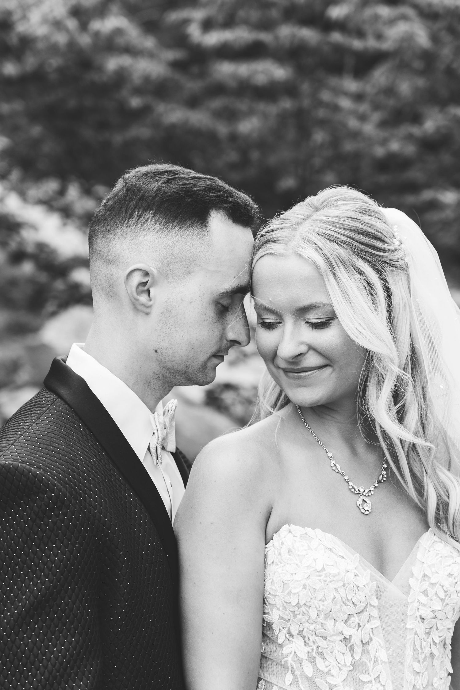 Black and white intimate wedding portrait of newlyweds sharing a tender moment outdoors with natural background.