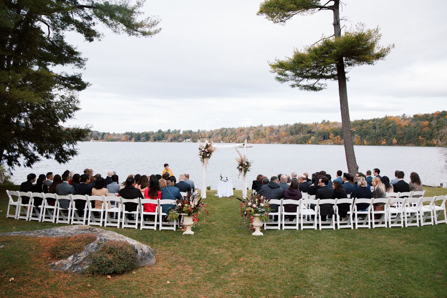 Outdoor lakeside wedding ceremony with white chairs set up on grass overlooking water and fall foliage.