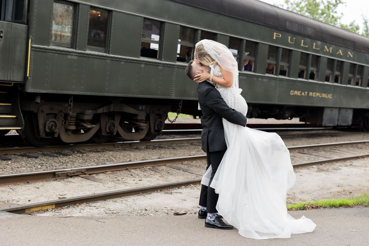 A romantic couple poses near vintage train cars for wedding photos, with the bride's white dress flowing in the breeze.