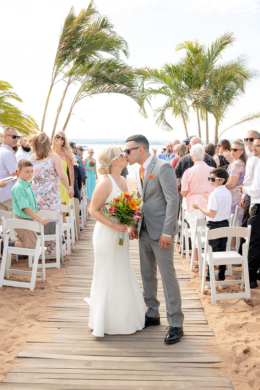 Beach wedding ceremony on wooden boardwalk with palm trees and guests seated in white chairs by the ocean.