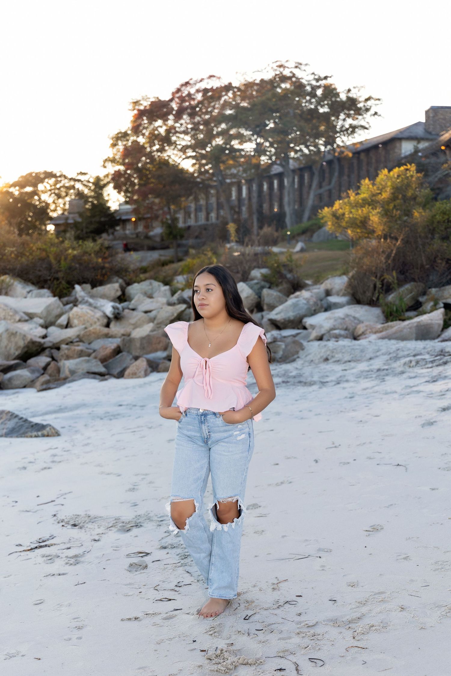 A woman in a pink top and light denim jeans poses casually on a sandy beach at sunset.