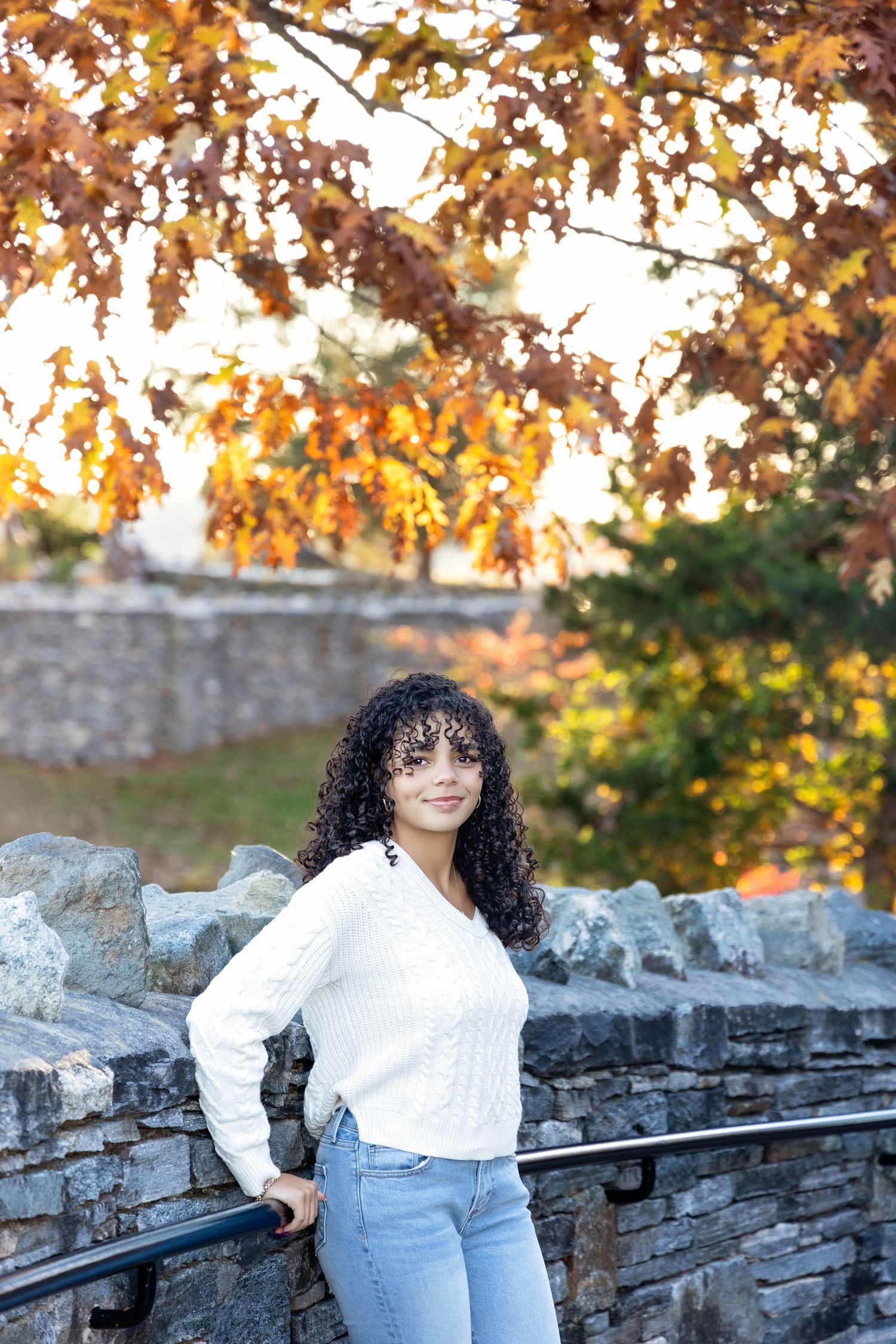 A person in casual attire poses by a stone wall during golden hour with fall foliage in background.