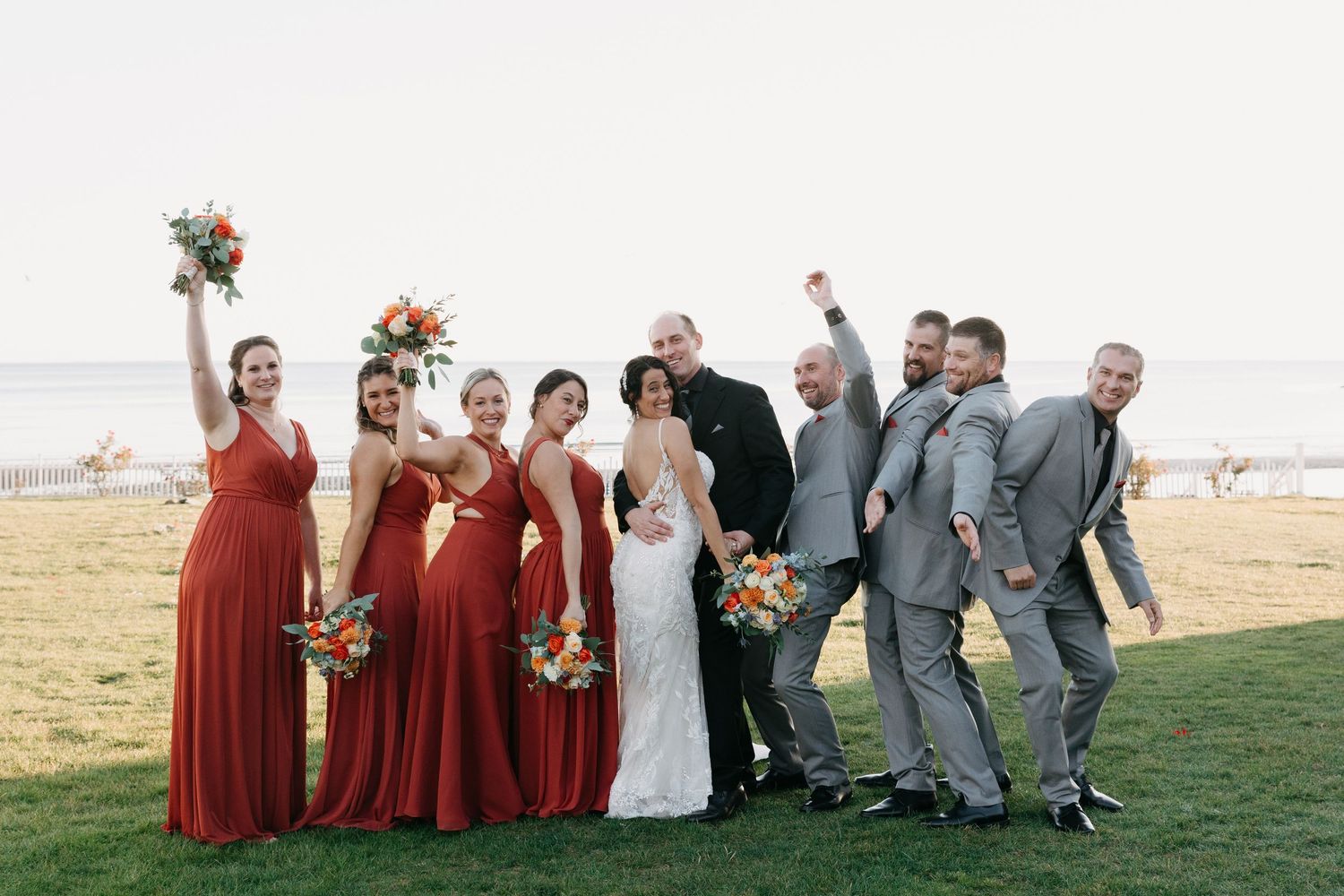 Wedding party in red bridesmaid dresses and gray suits poses joyfully on coastal lawn.