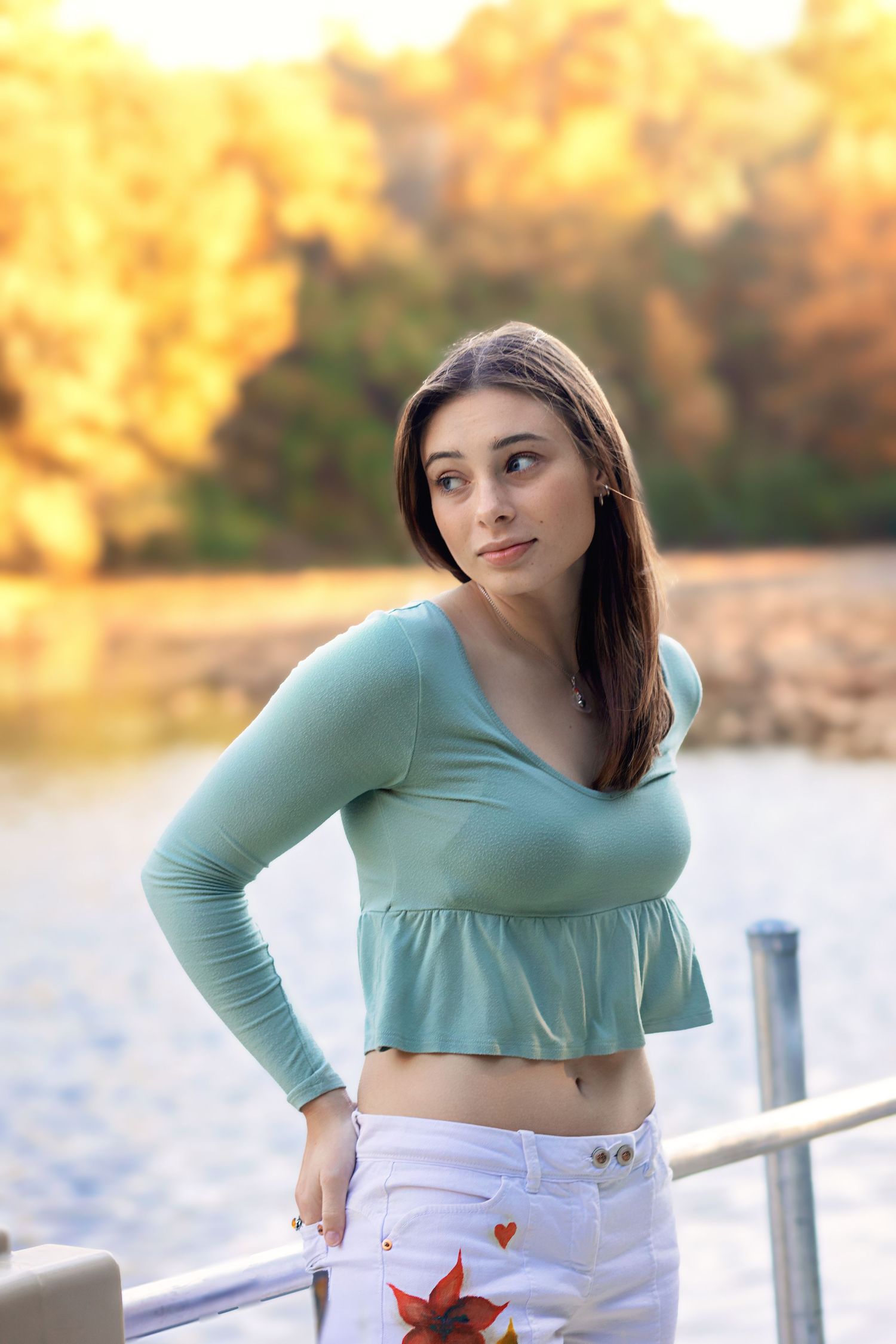 A person in a mint green cropped top and white pants poses by a lake during golden hour with autumn foliage in background.