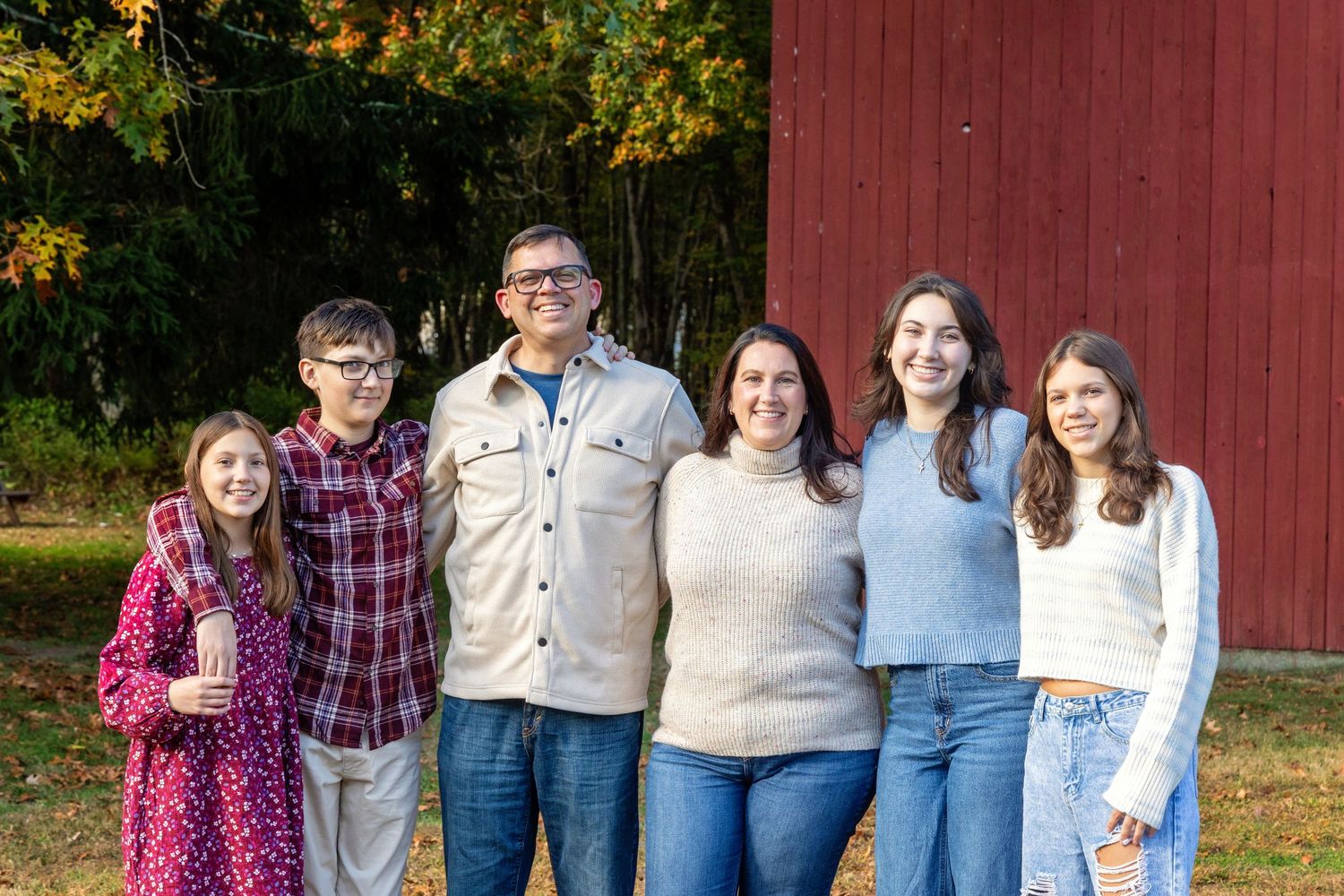 A group stands together in casual attire against a red barn wall on an autumn day.