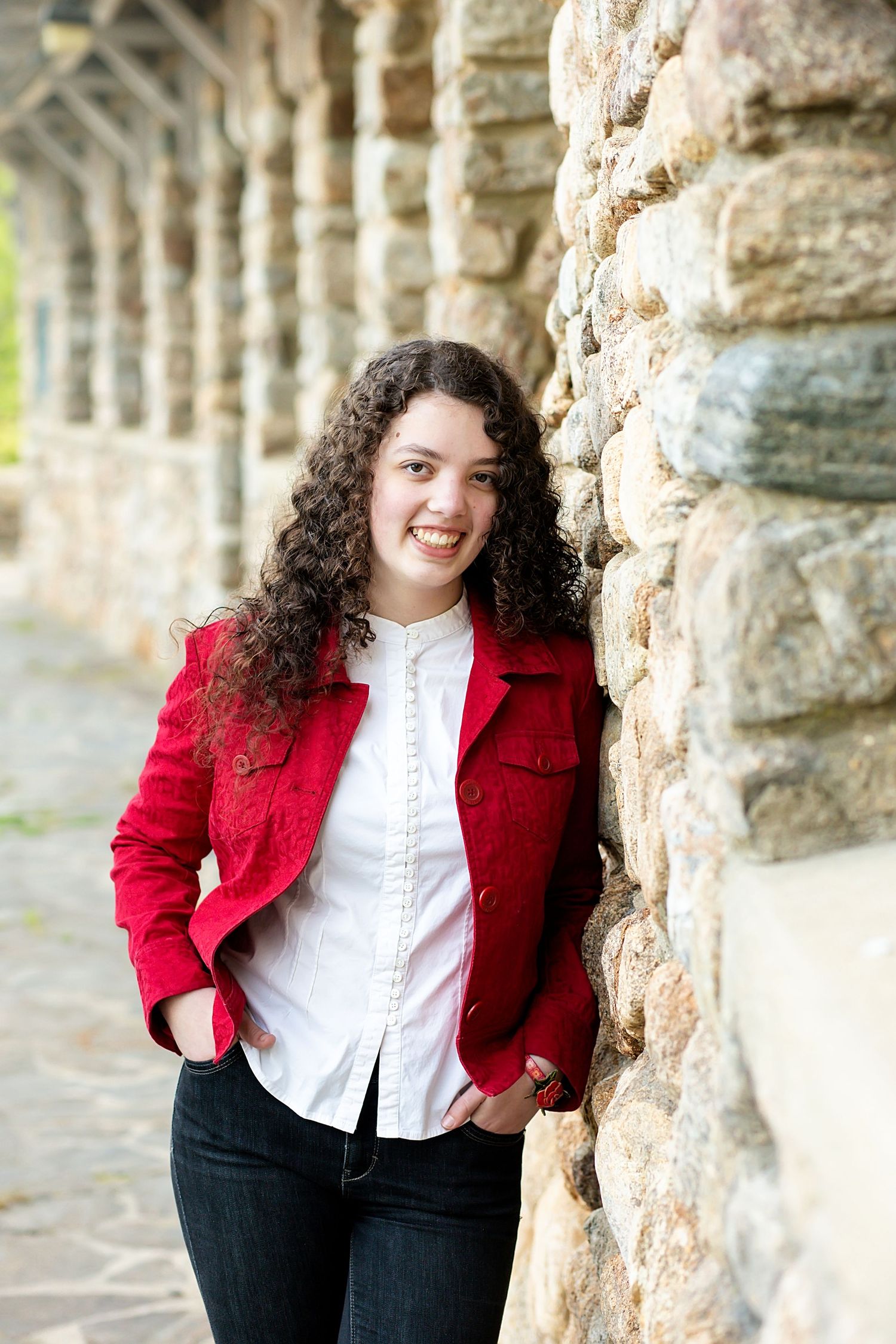 A person in a red blazer and white button-up shirt stands against a stone wall in an outdoor colonnade.