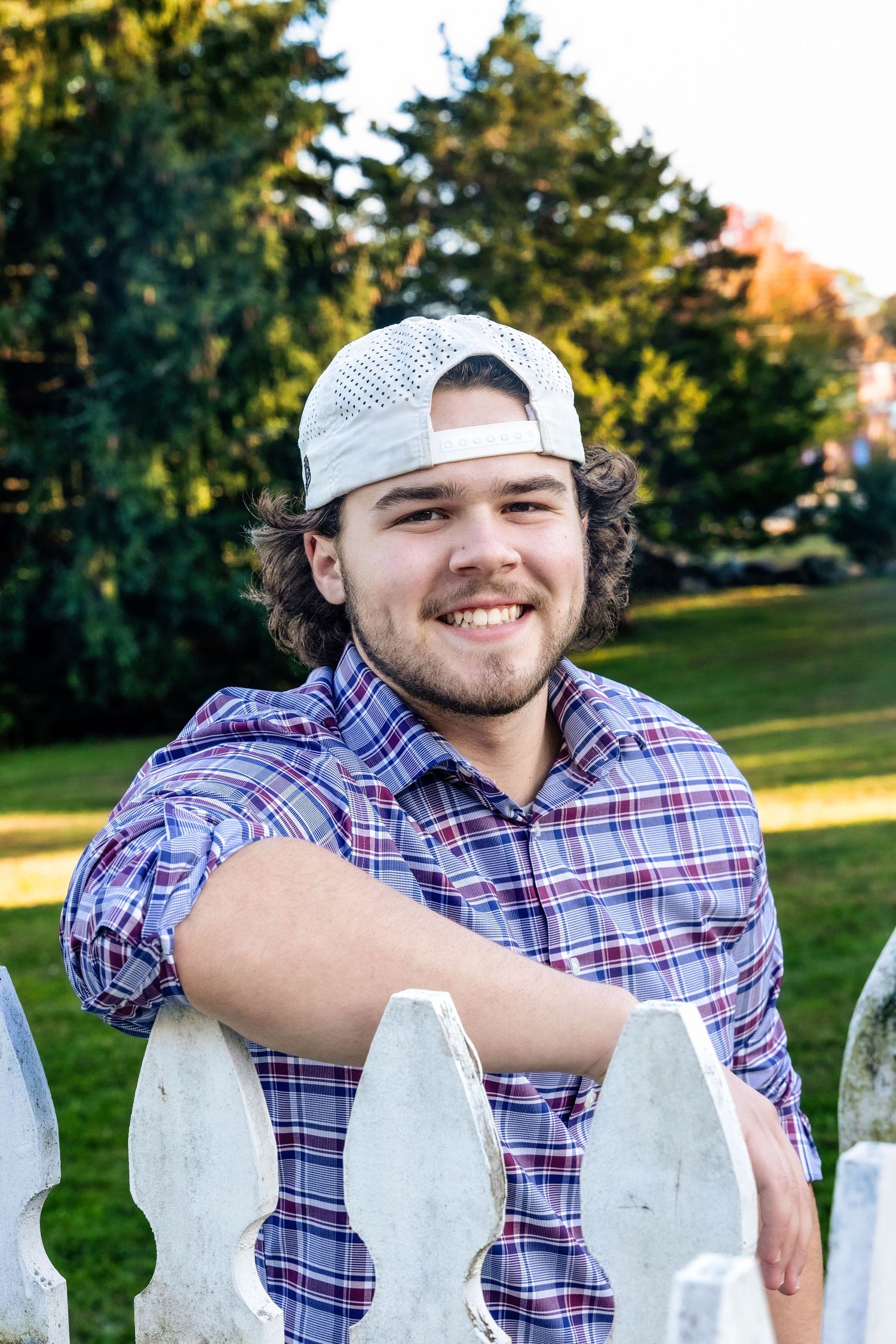 A person in a plaid shirt and baseball cap leans on a white picket fence in a sunny outdoor setting.