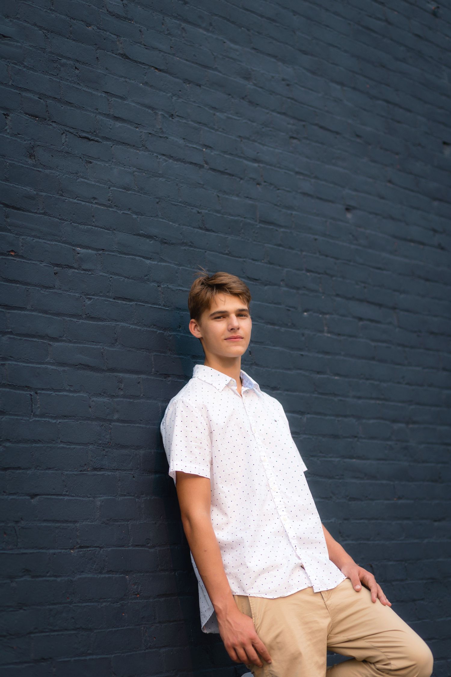 A person in a white polka dot shirt and khaki pants leans against a dark gray brick wall.