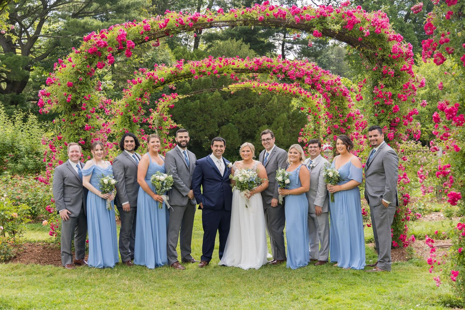 Wedding party poses under vibrant pink bougainvillea arch in garden setting with light blue bridesmaid dresses.